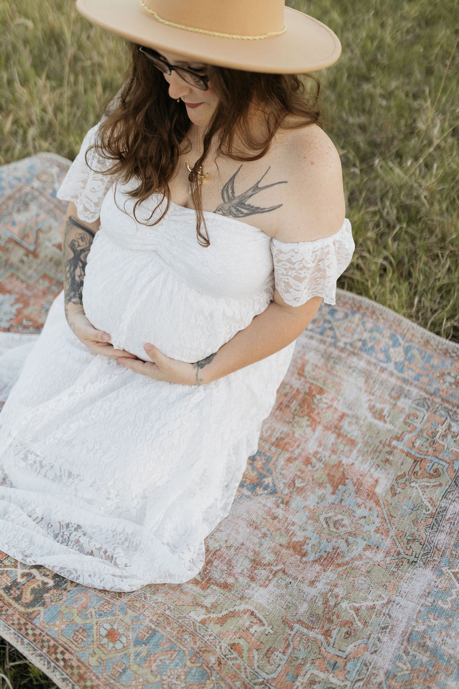 An expectant mother in a white lace dress sits on a patterned blanket in a grassy field.
