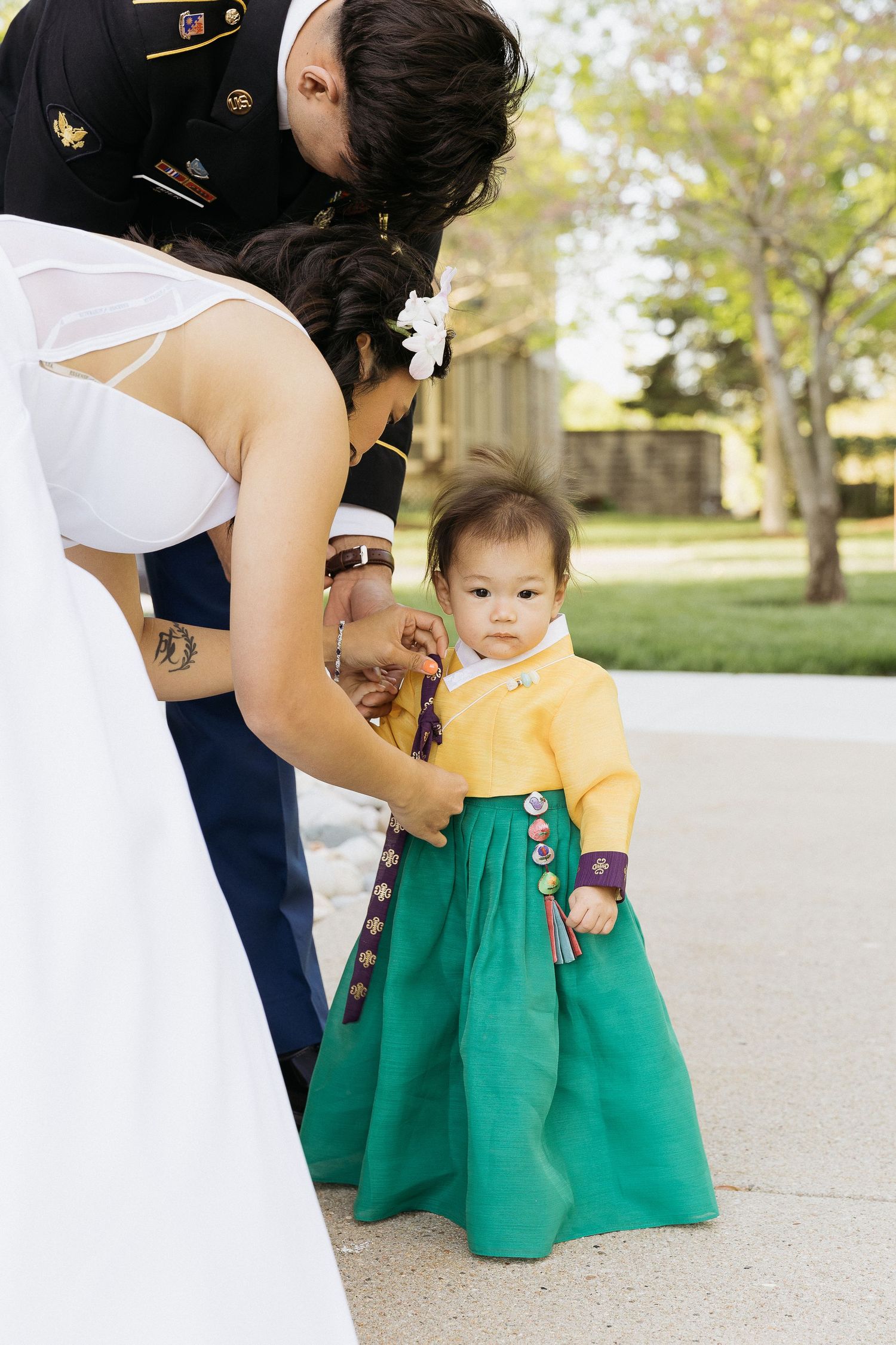 A baby dressed in yellow and green stands beside adults in formal attire during a wedding celebration.