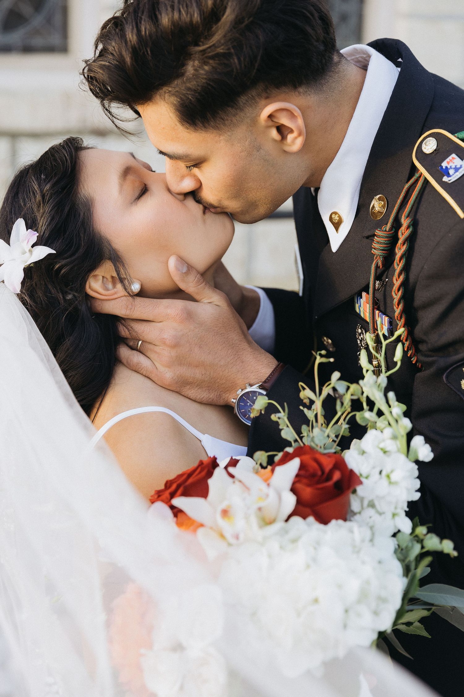 A romantic wedding portrait shows a couple kissing as the bride holds a bouquet with red and white flowers.