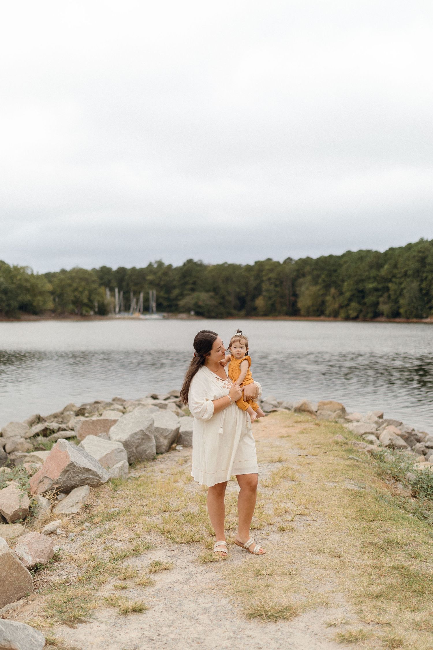 Mother and child stand on rocky peninsula overlooking peaceful lake.