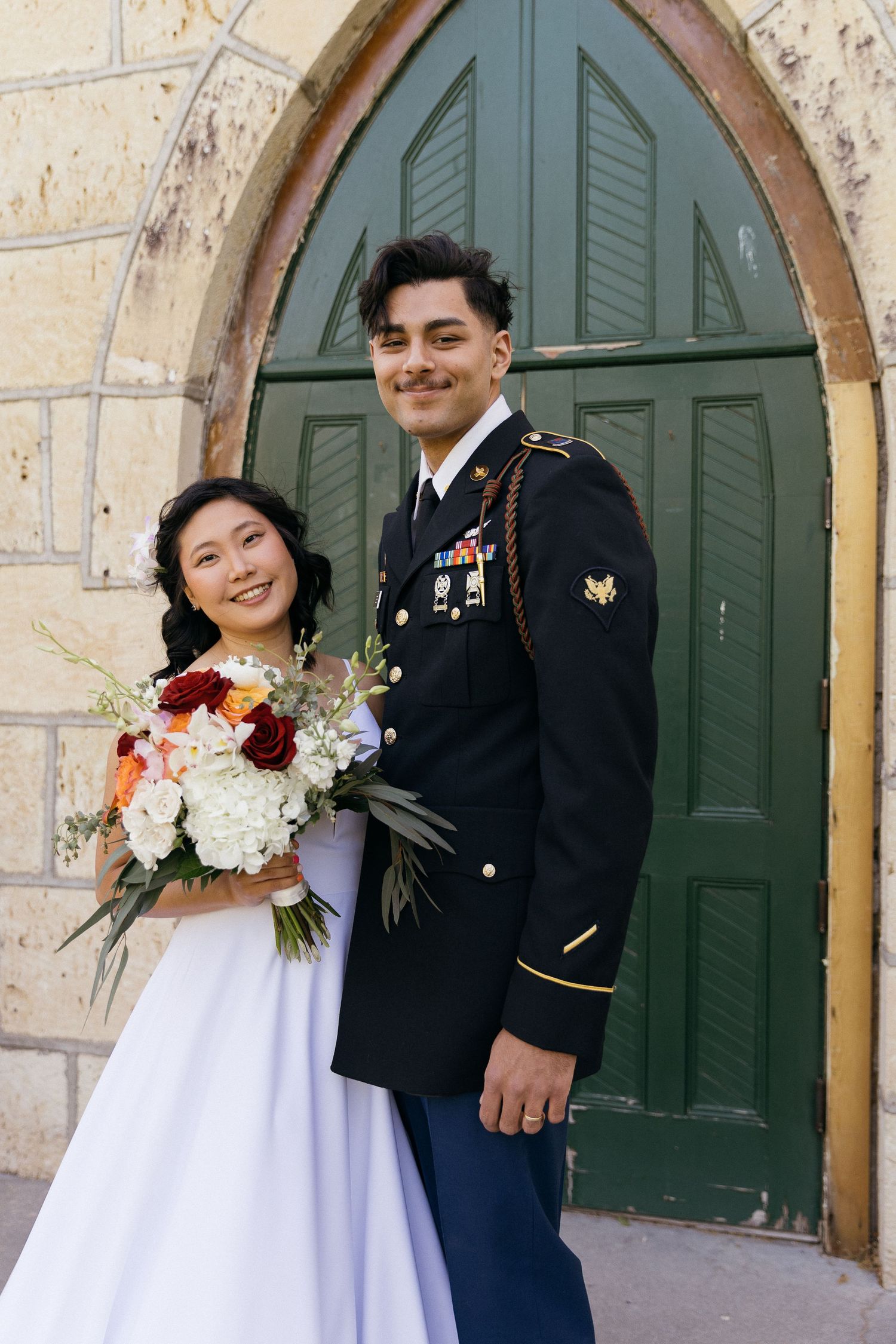 A wedding couple poses in front of an arched green door at a stone church.