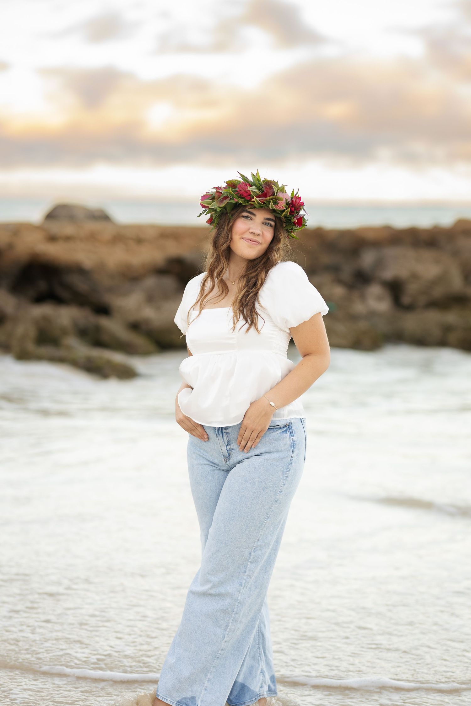 Expectant mother in white shirt and jeans wears floral crown on beach at sunset.