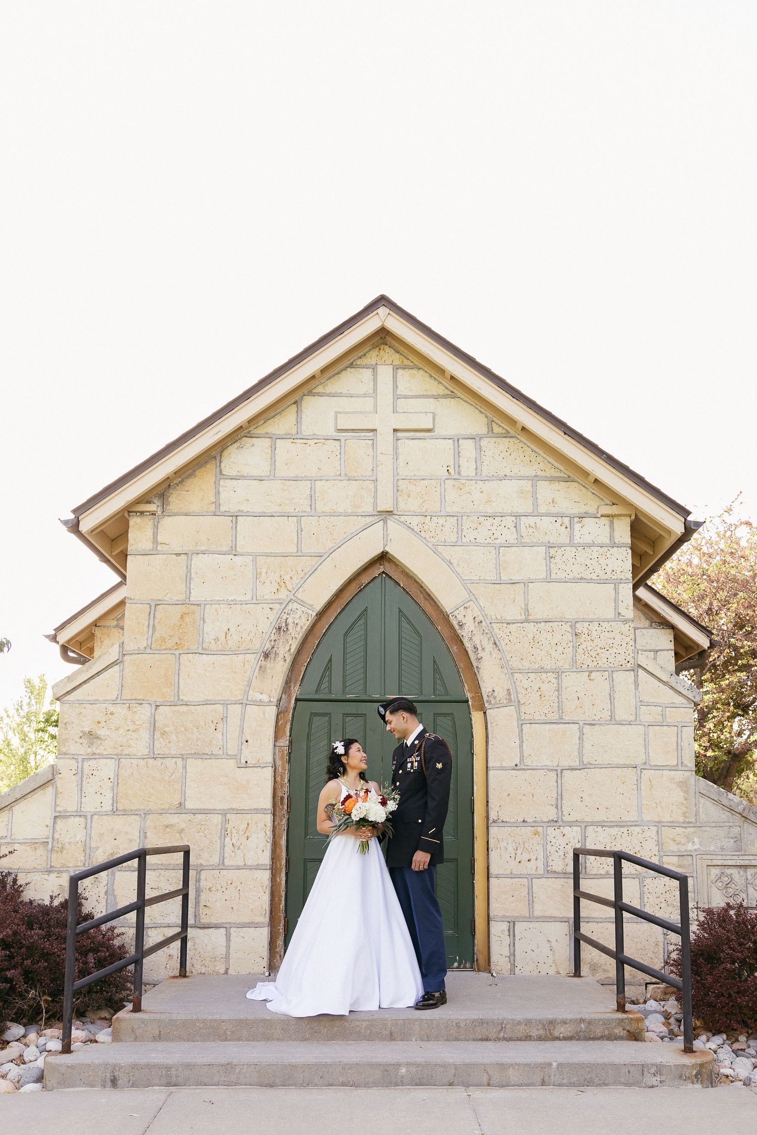 A wedding couple poses in front of a small stone chapel with a cross above the entrance.