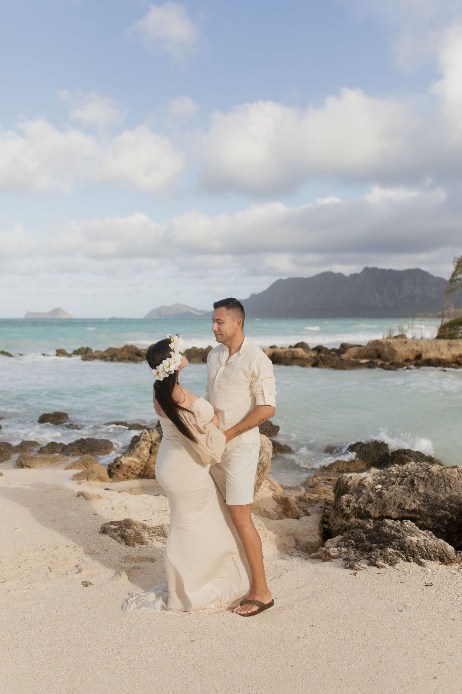 A couple embracing on a beach with rocky outcrops and mountains in the distance.