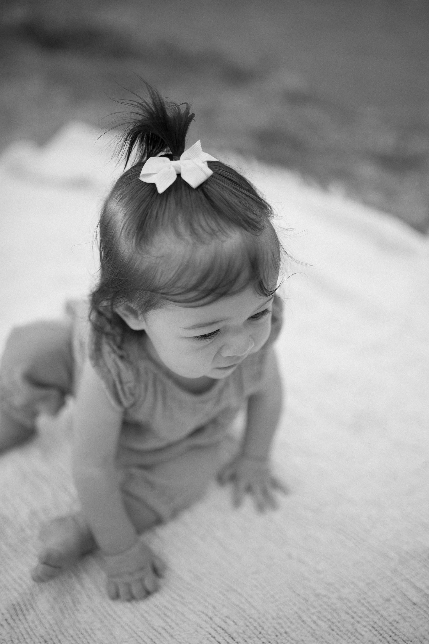 Black and white portrait of toddler with bow sitting on blanket.