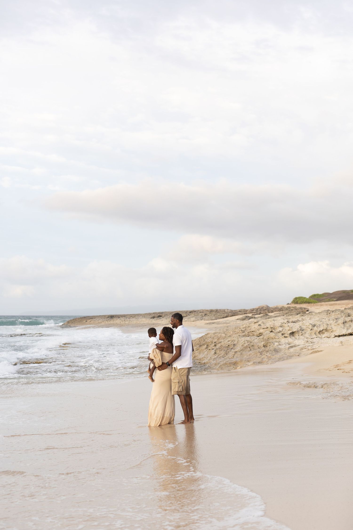 A couple stands together on a serene beach at sunset with waves lapping at their feet and sandy dunes in the distance.