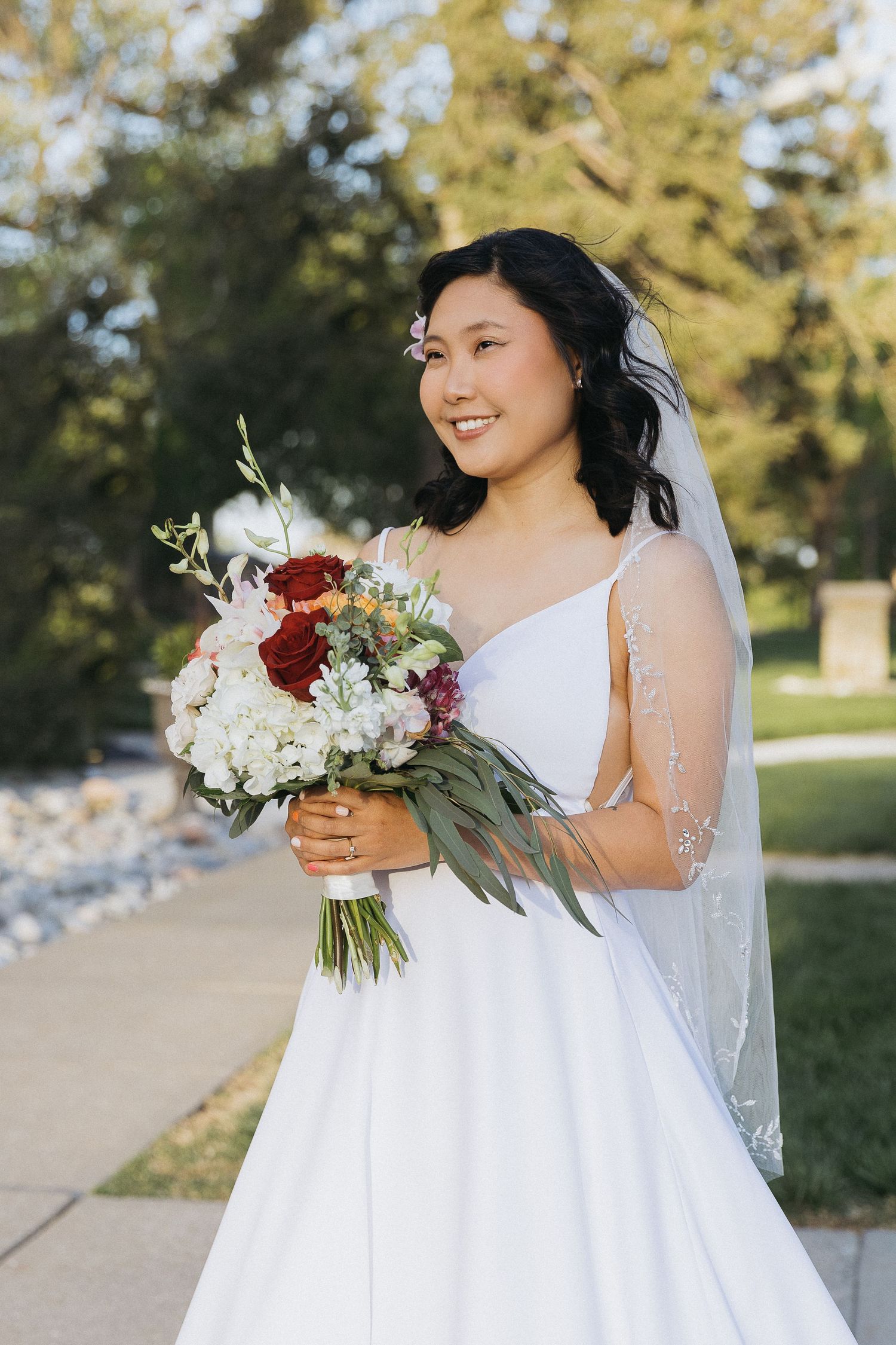 A bride wearing a white gown holds a red and white bouquet while smiling outdoors.