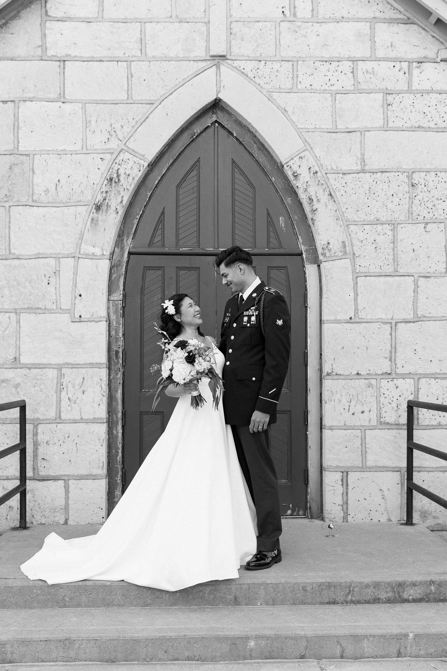 A wedding couple stands in front of a gothic arched church door in a black and white photo.