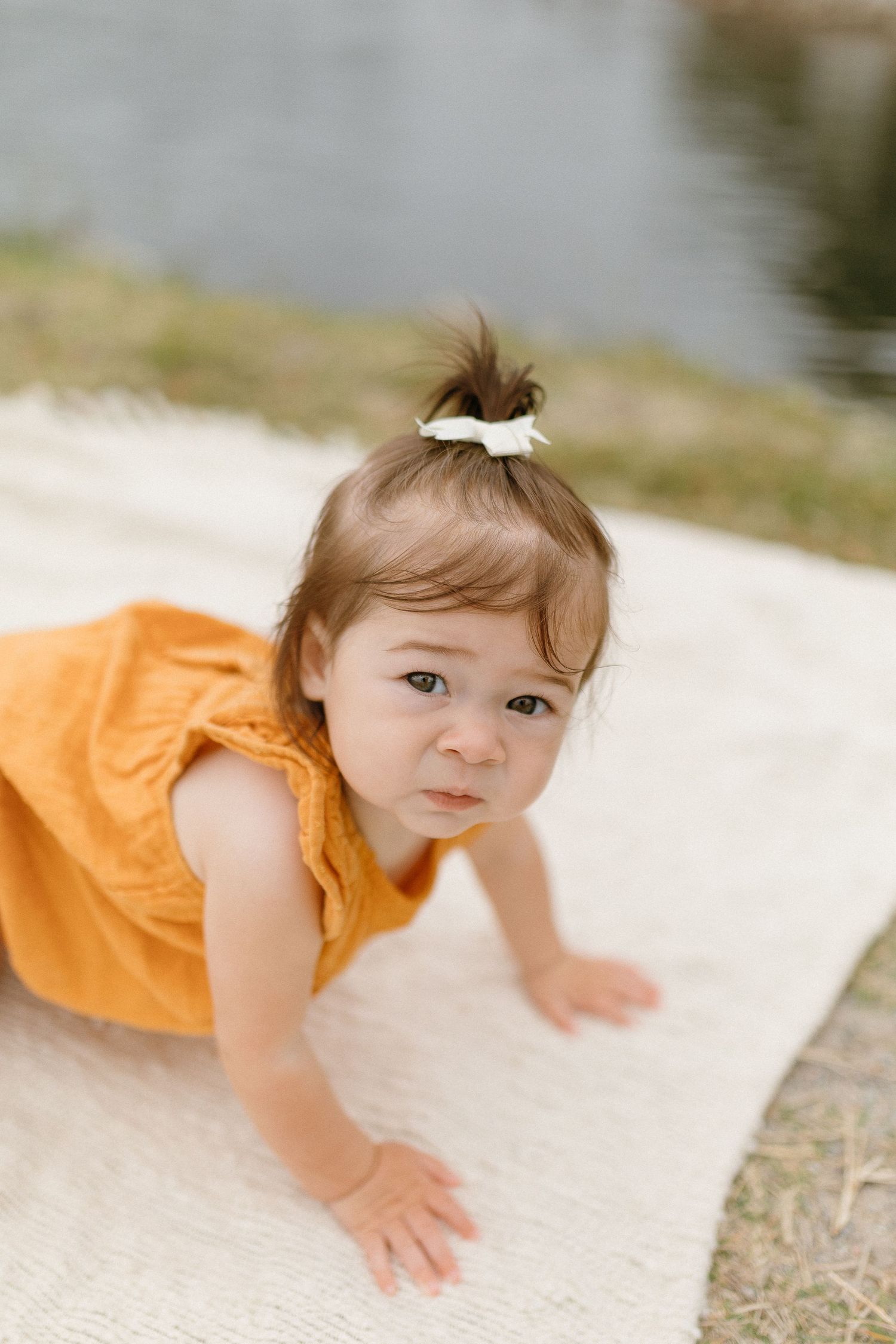 Close up of toddler in orange dress crawling on white blanket outdoors.