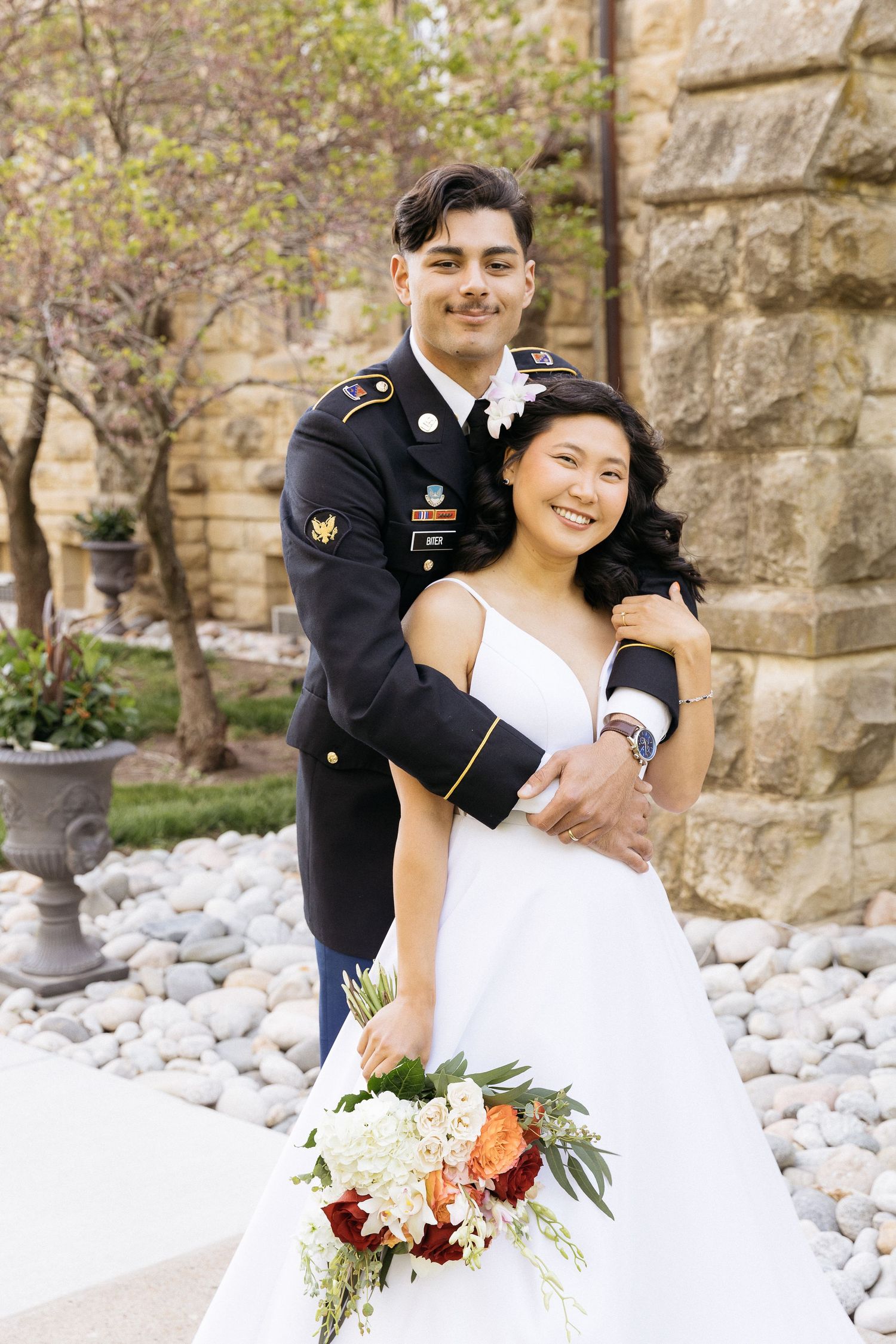 Military wedding couple smiling and embracing outdoors with stone wall and decorative florals in romantic outdoor setting.