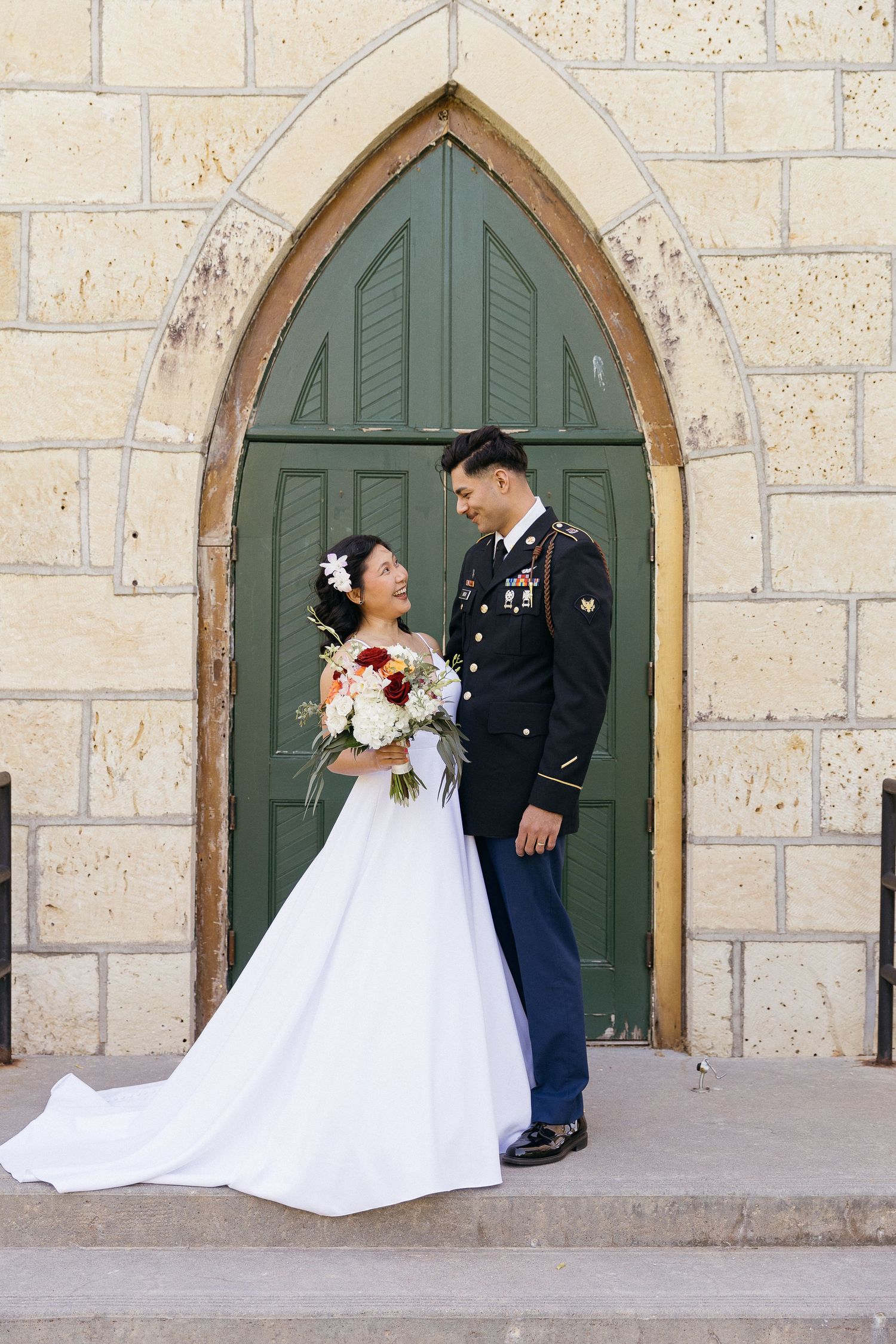 A military wedding couple shares a romantic moment in front of a gothic church doorway.