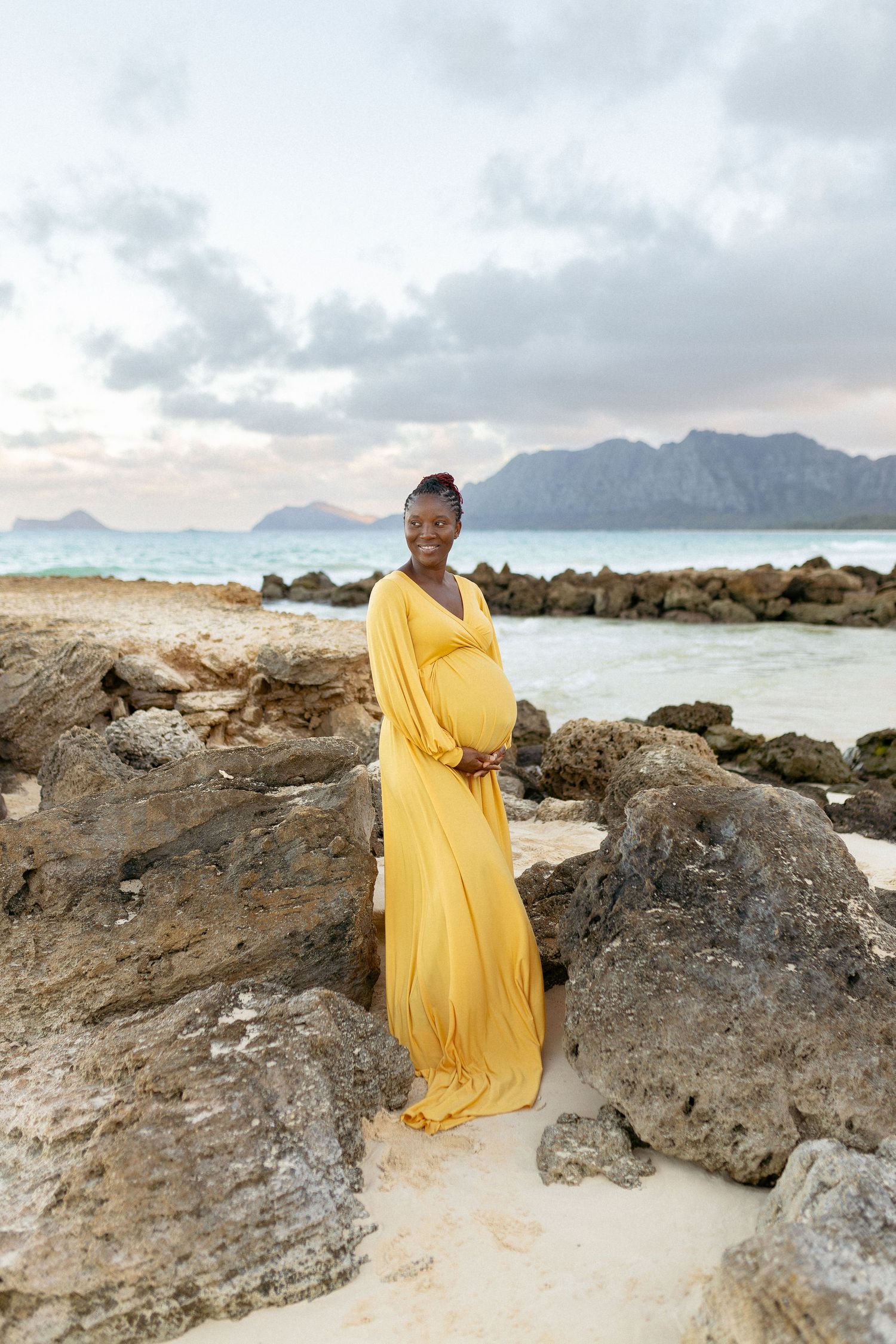 A woman in a yellow flowing dress stands among coastal rocks during sunset.