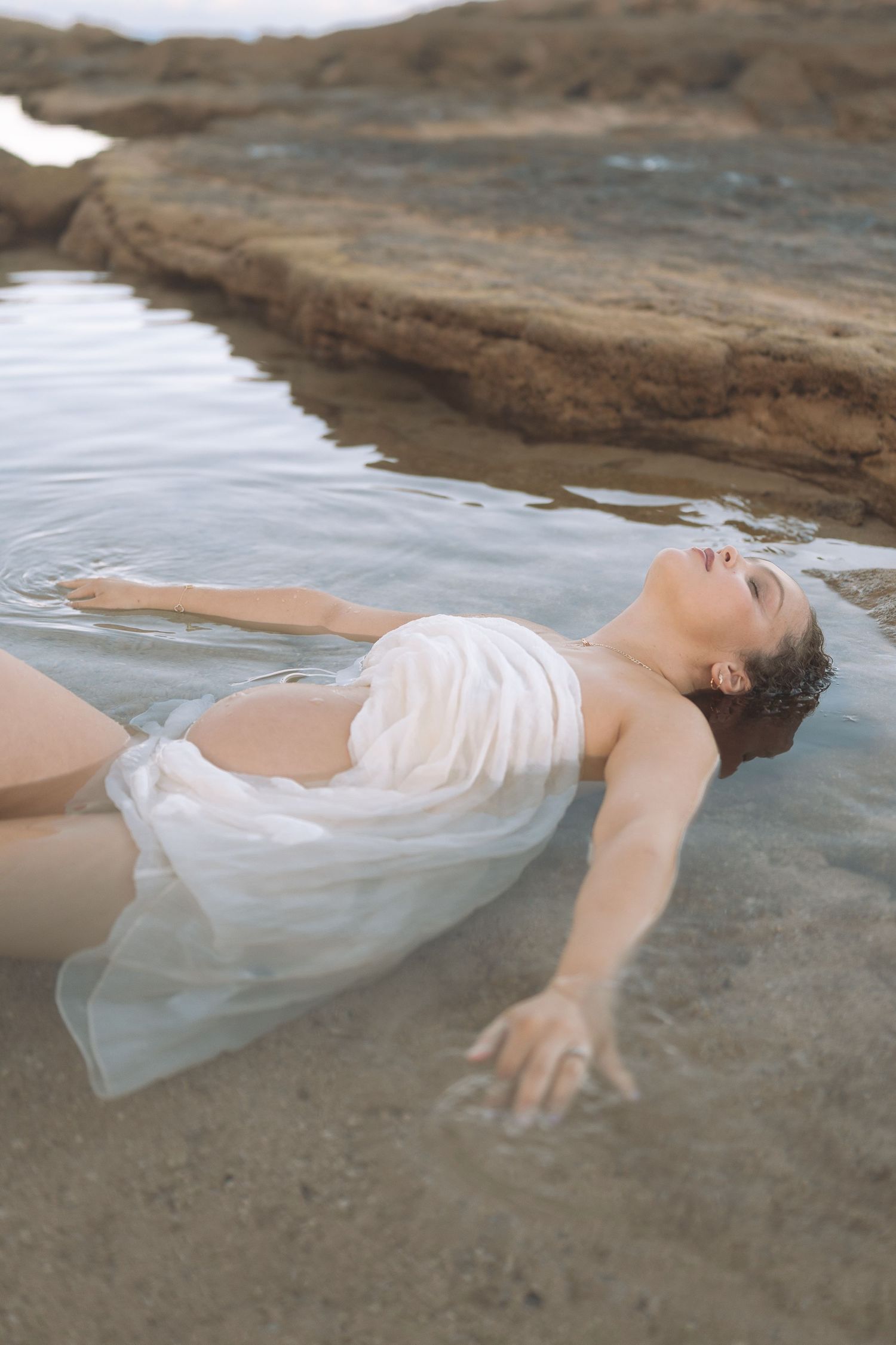 A figure in flowing white fabric lies gracefully on rocks near water in an ethereal artistic pose.
