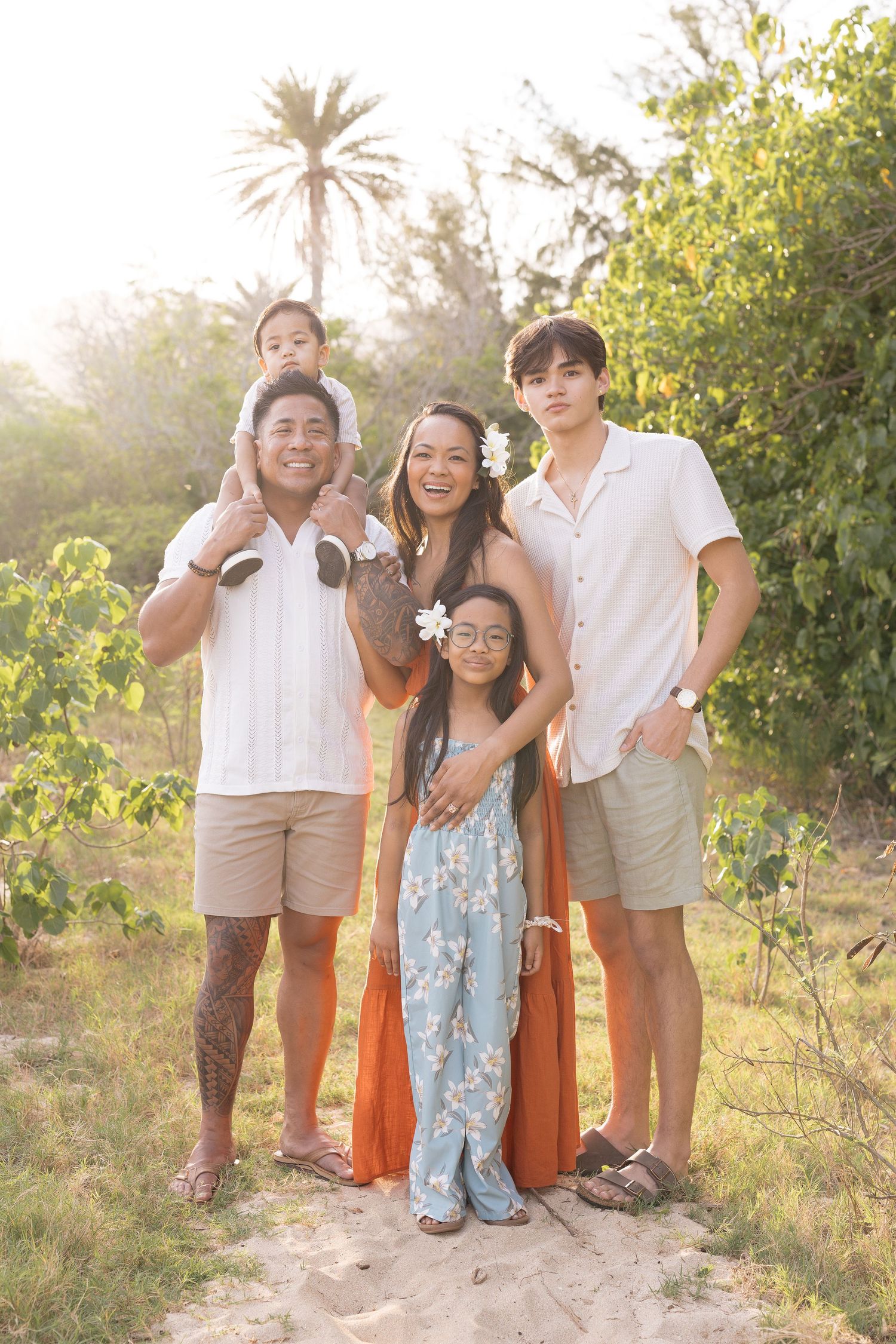 A family poses together outdoors at golden hour with a palm tree and lush greenery in the background.