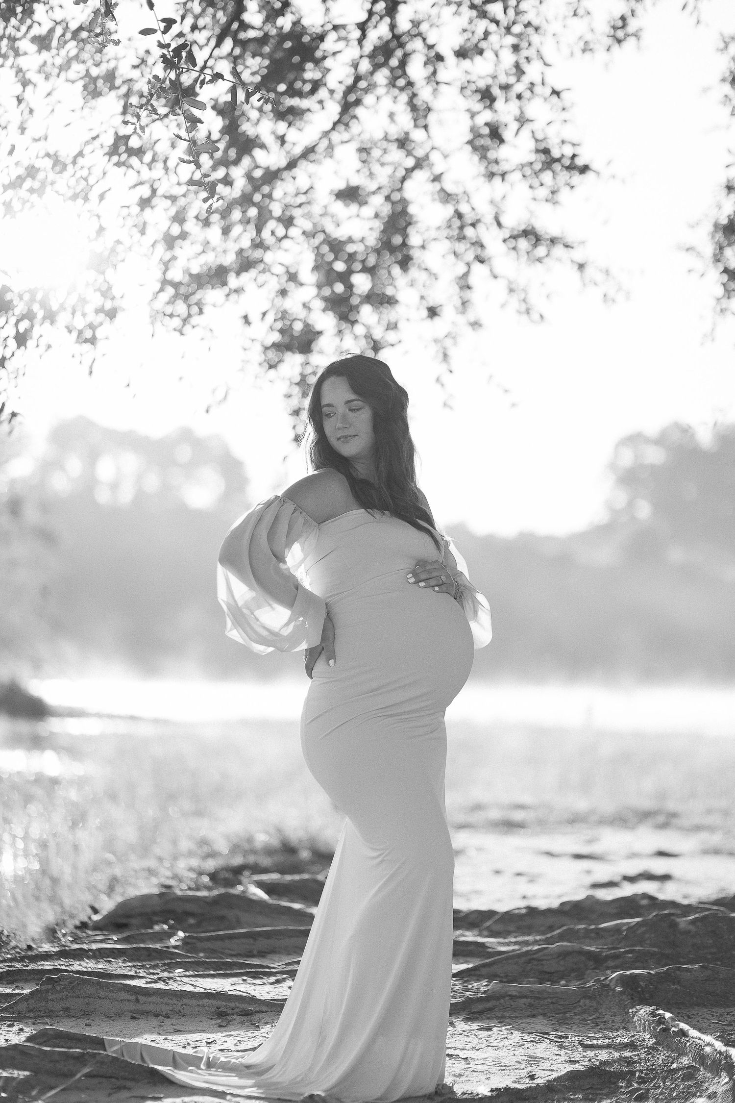 Artistic black and white maternity photo of a figure in a flowing dress standing near water at sunset.