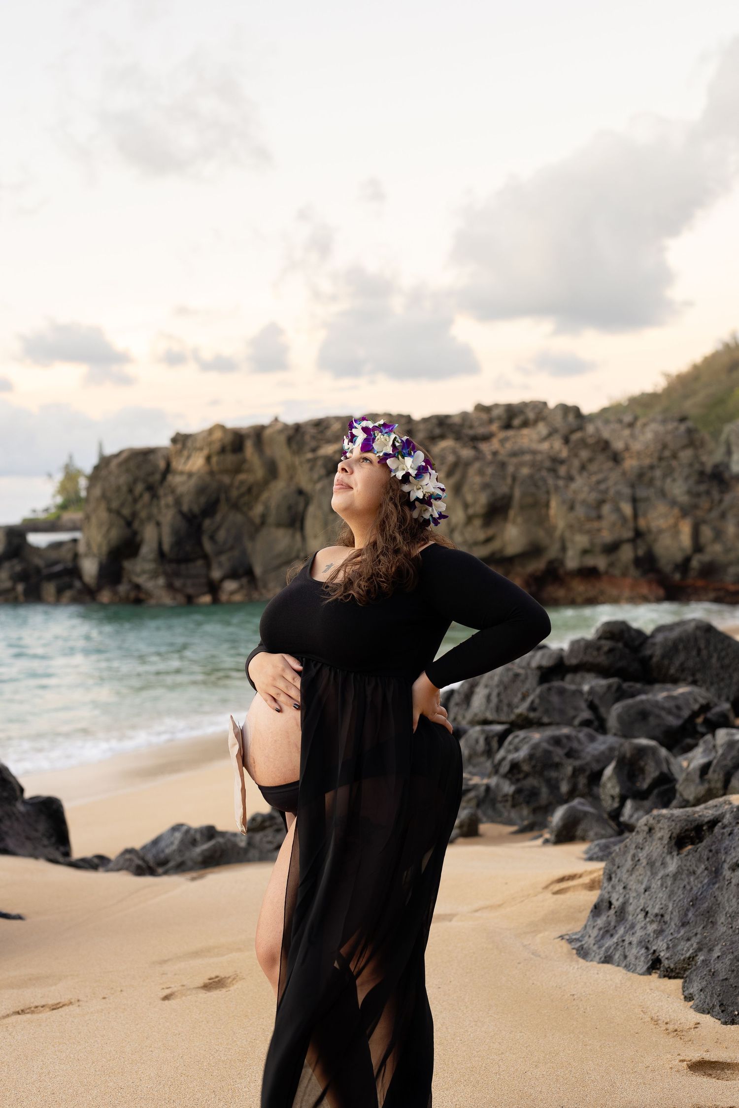 A maternity portrait on rocky beach with woman in black dress wearing floral crown.
