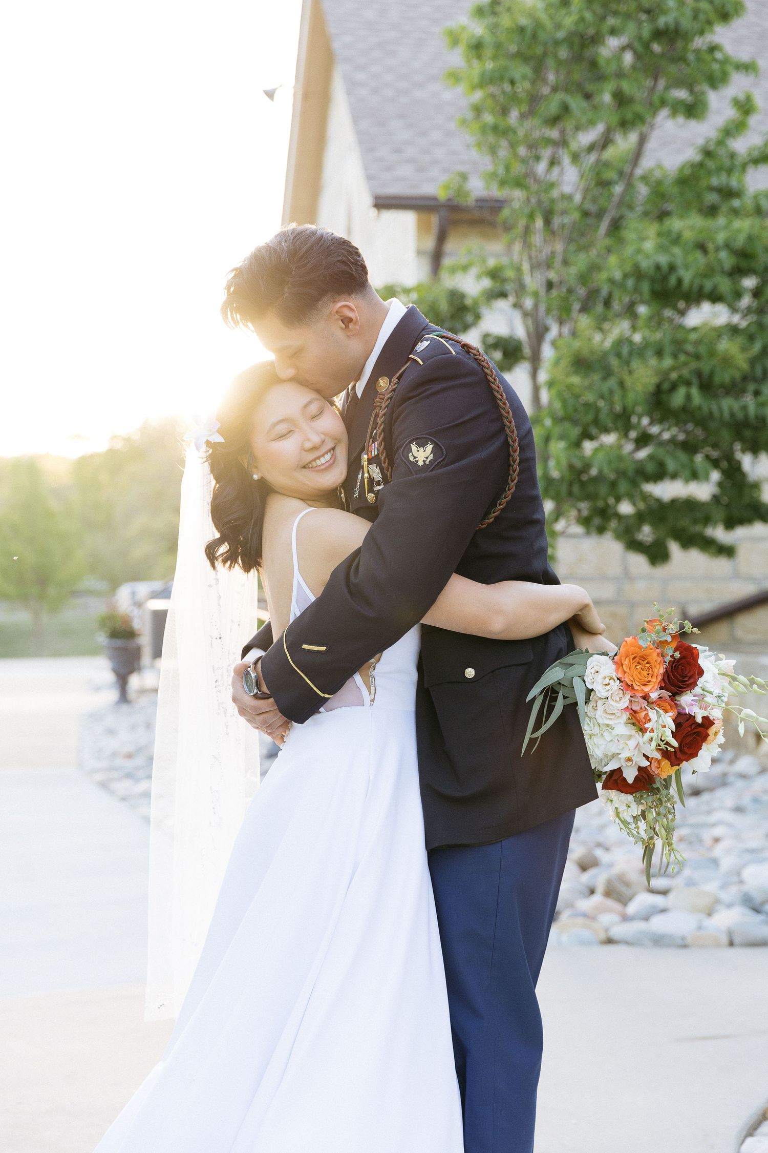 A newlywed couple embraces in warm sunlight as the bride holds an autumn-colored bouquet beside a stone wall.