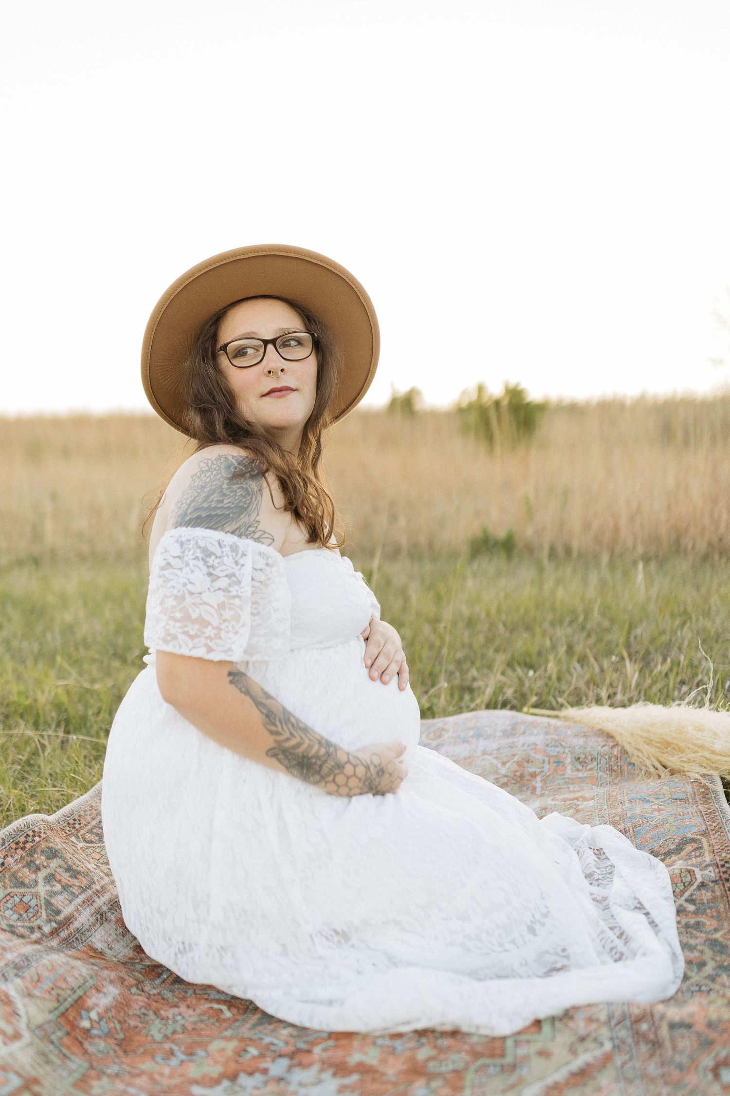 Maternity photo on a blanket featuring white lace dress and brown hat in a grassy field.