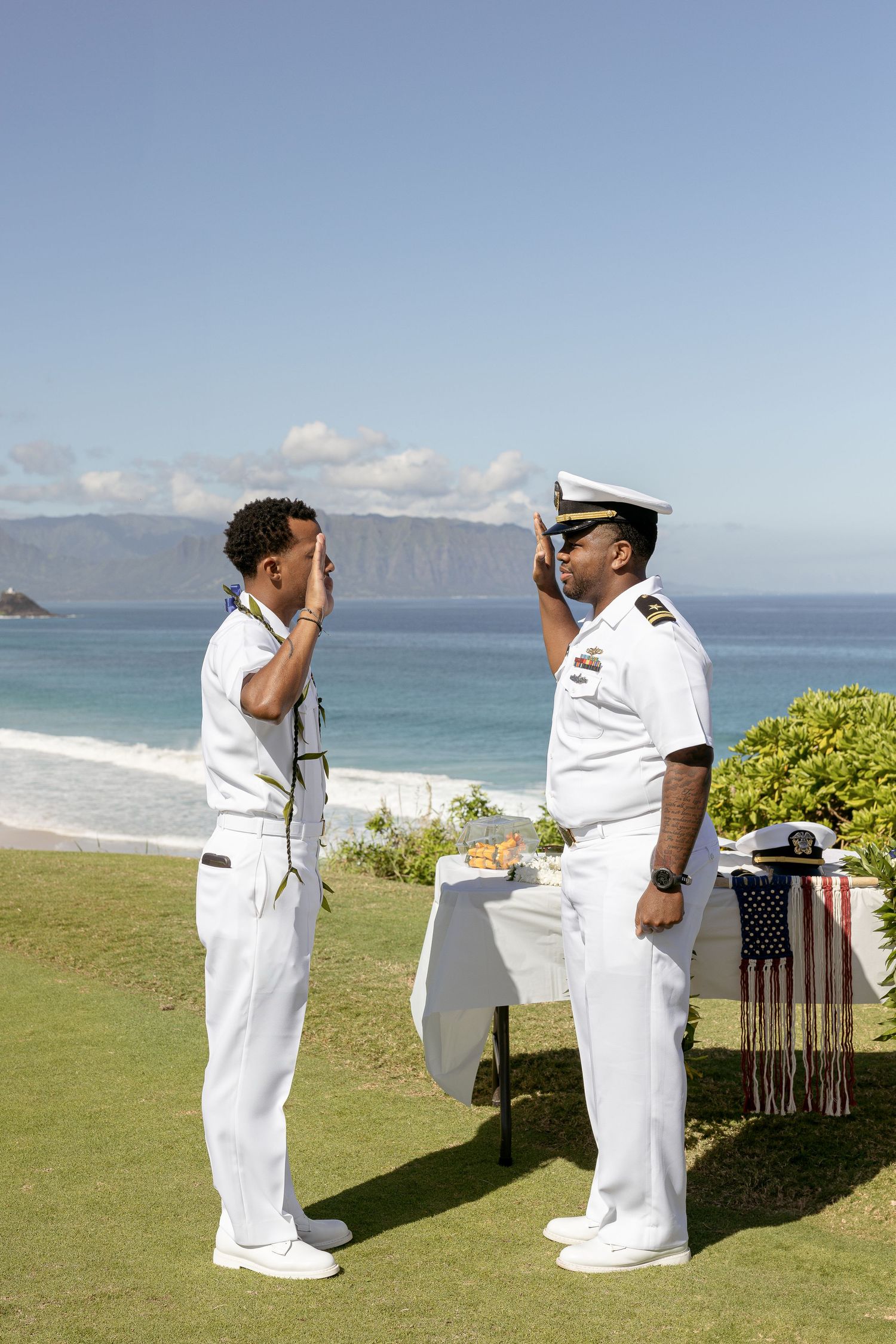 Navy officers in white uniforms stand on a grassy cliff overlooking a scenic beach and ocean view.