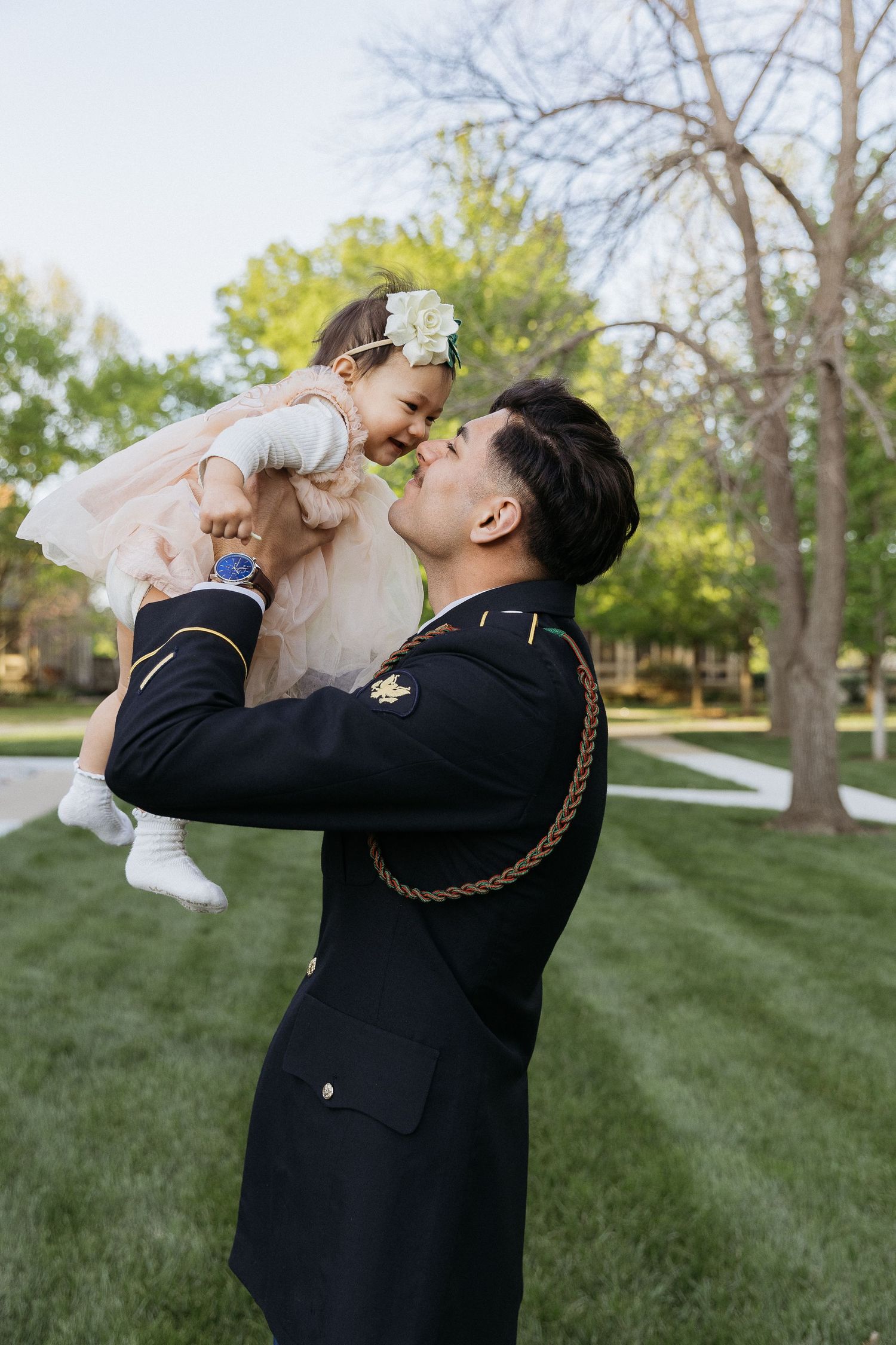 Military father playfully lifts young daughter in dress on grassy lawn.
