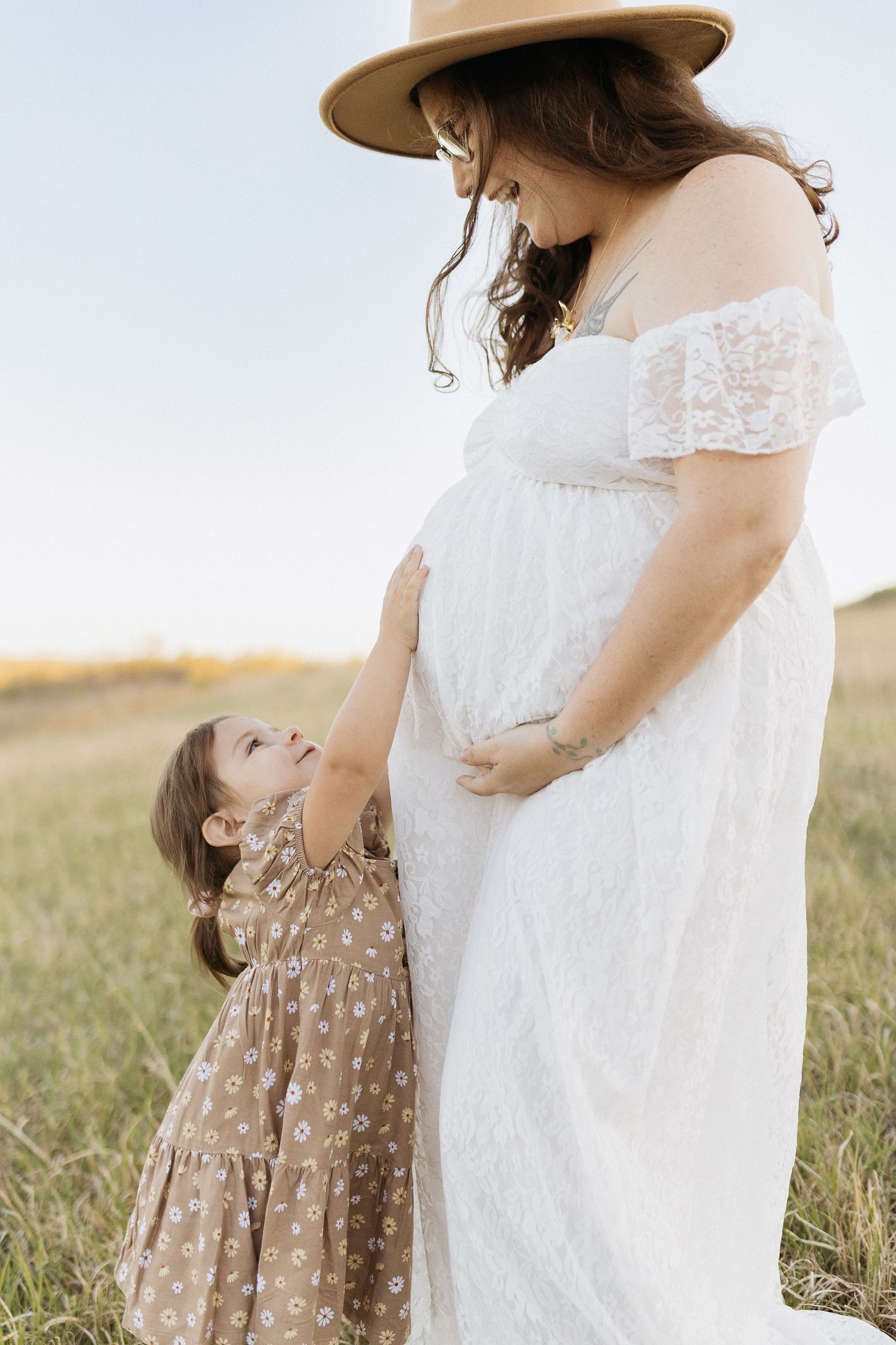 Sweet maternity moment with small child reaching up to touch white lace dress in sunset field.