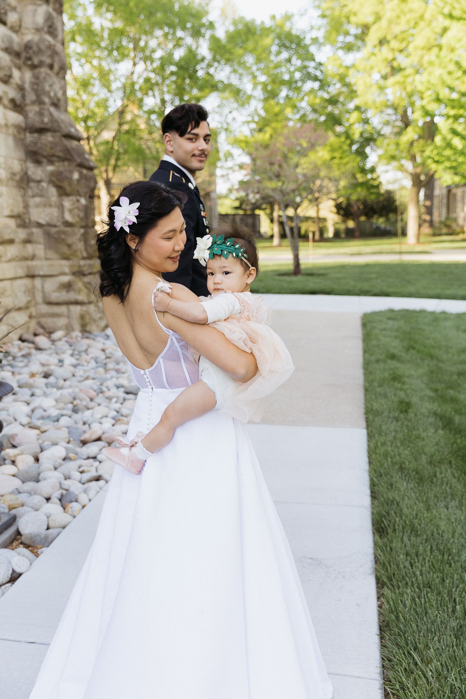 Wedding couple poses with baby in white dress and military uniform near stone building.