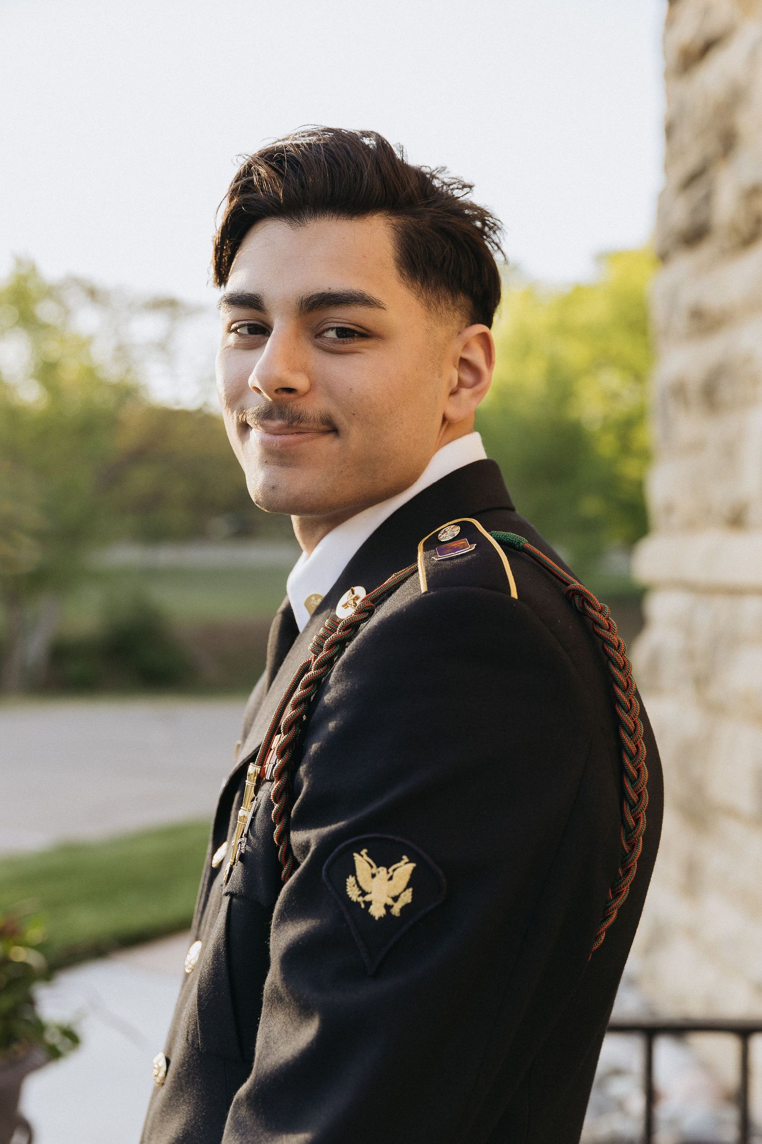 A military service member in dress uniform poses in front of a stone building.