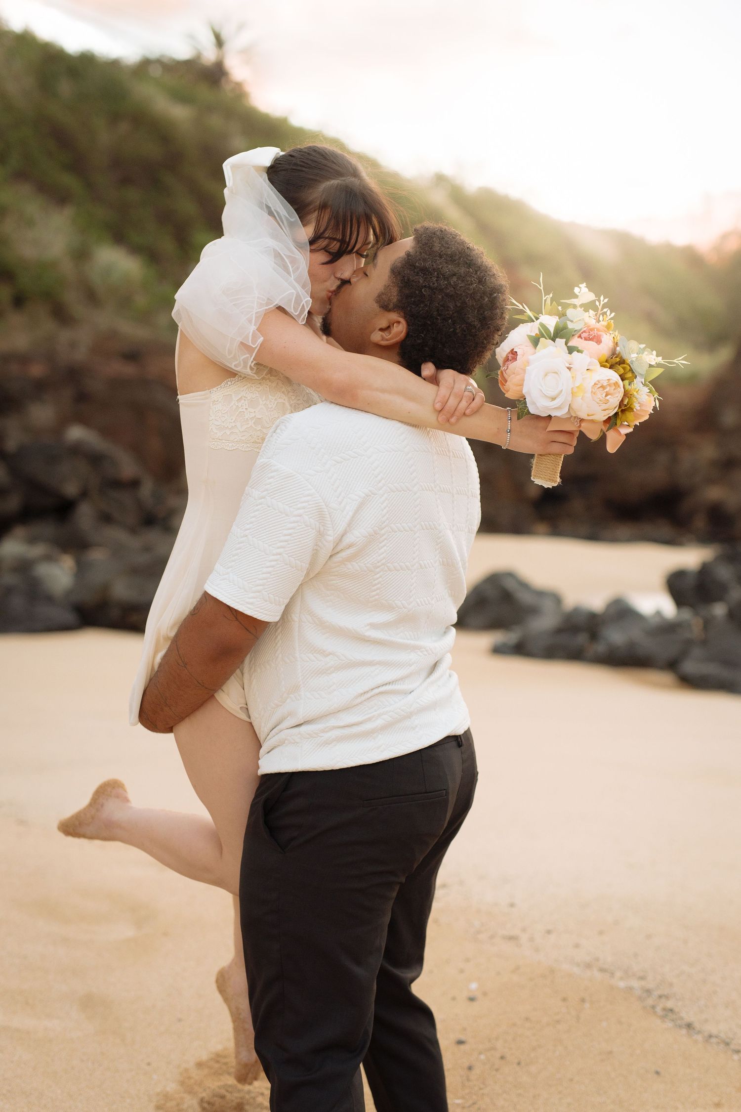 A romantic beach wedding moment with couple sharing embrace while holding white floral bouquet at sunset.