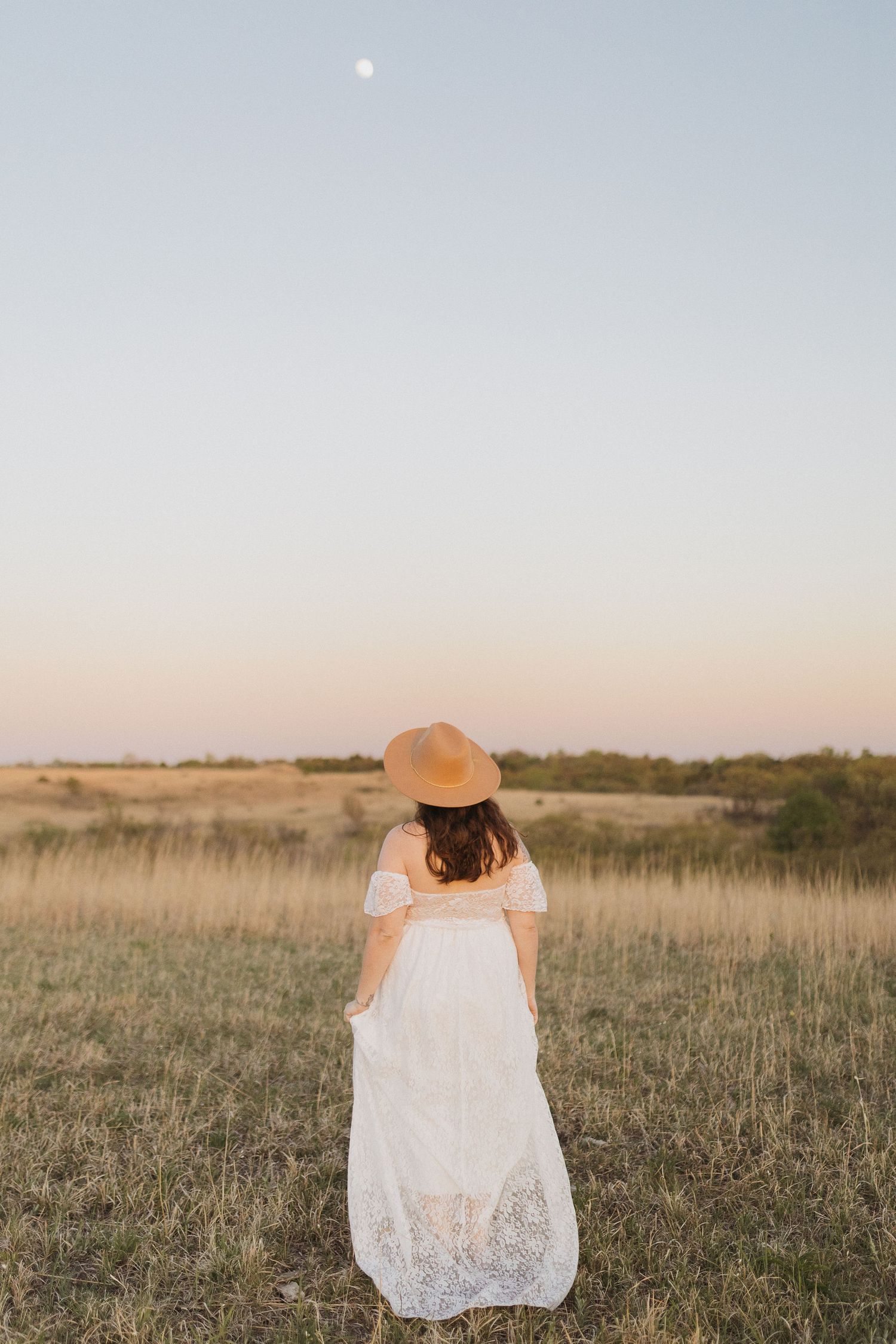 A figure in a white lace dress and brown hat stands in a golden field at sunset with the moon visible.