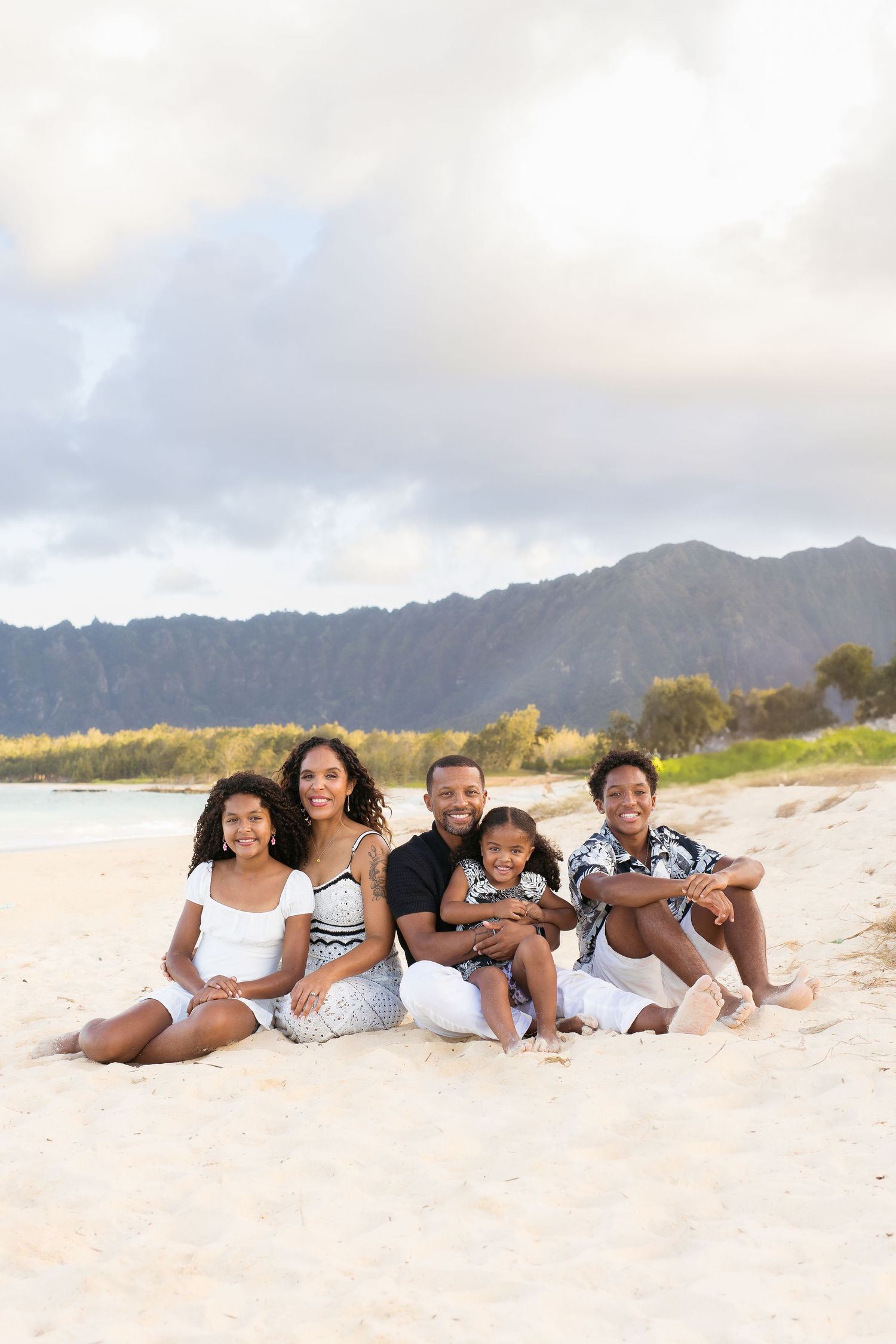 A happy family sits together on a sandy beach with mountains in the background during golden hour.