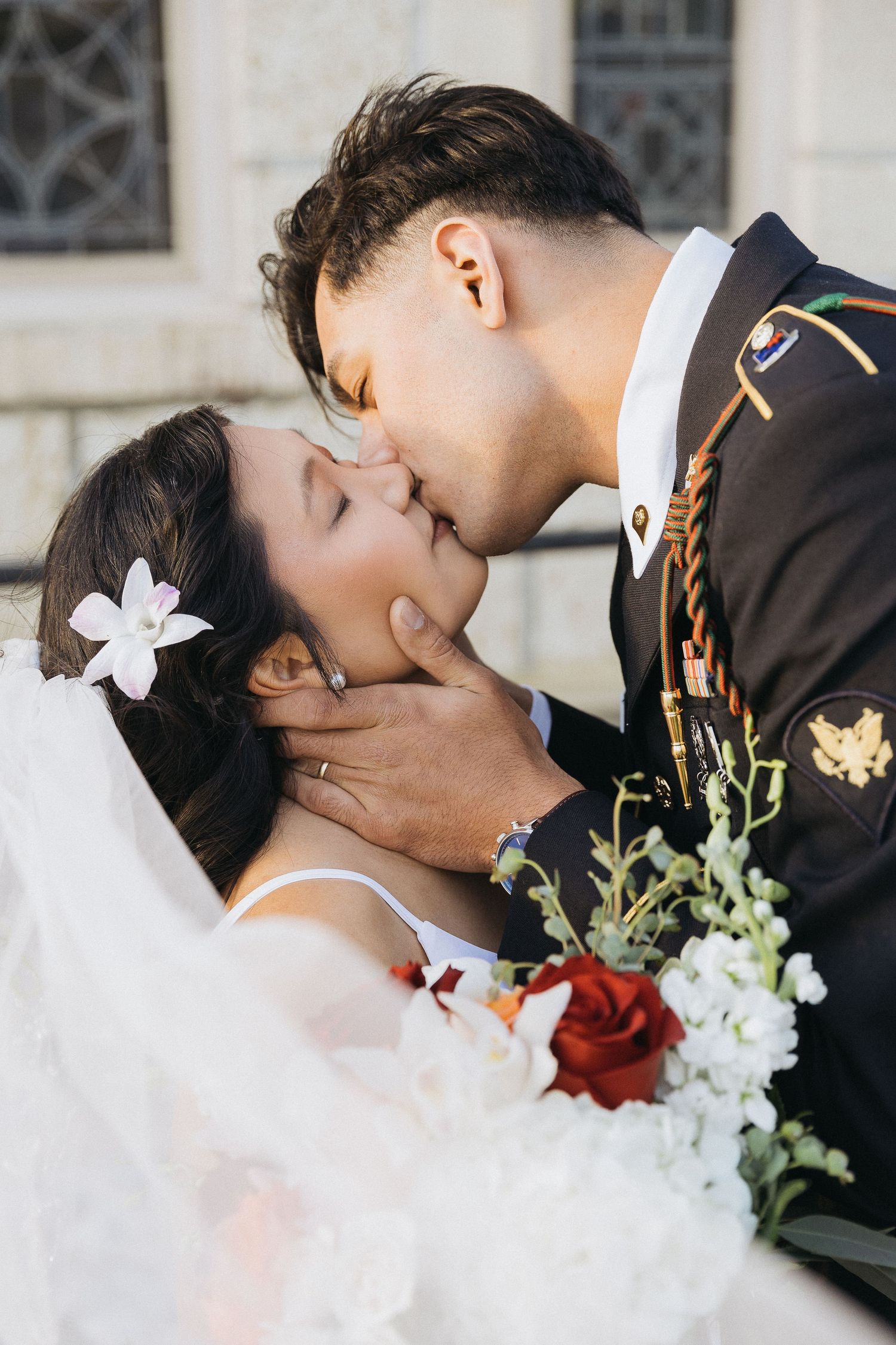 A romantic wedding kiss moment with red and white florals, white veil, and ornate architectural details in background.