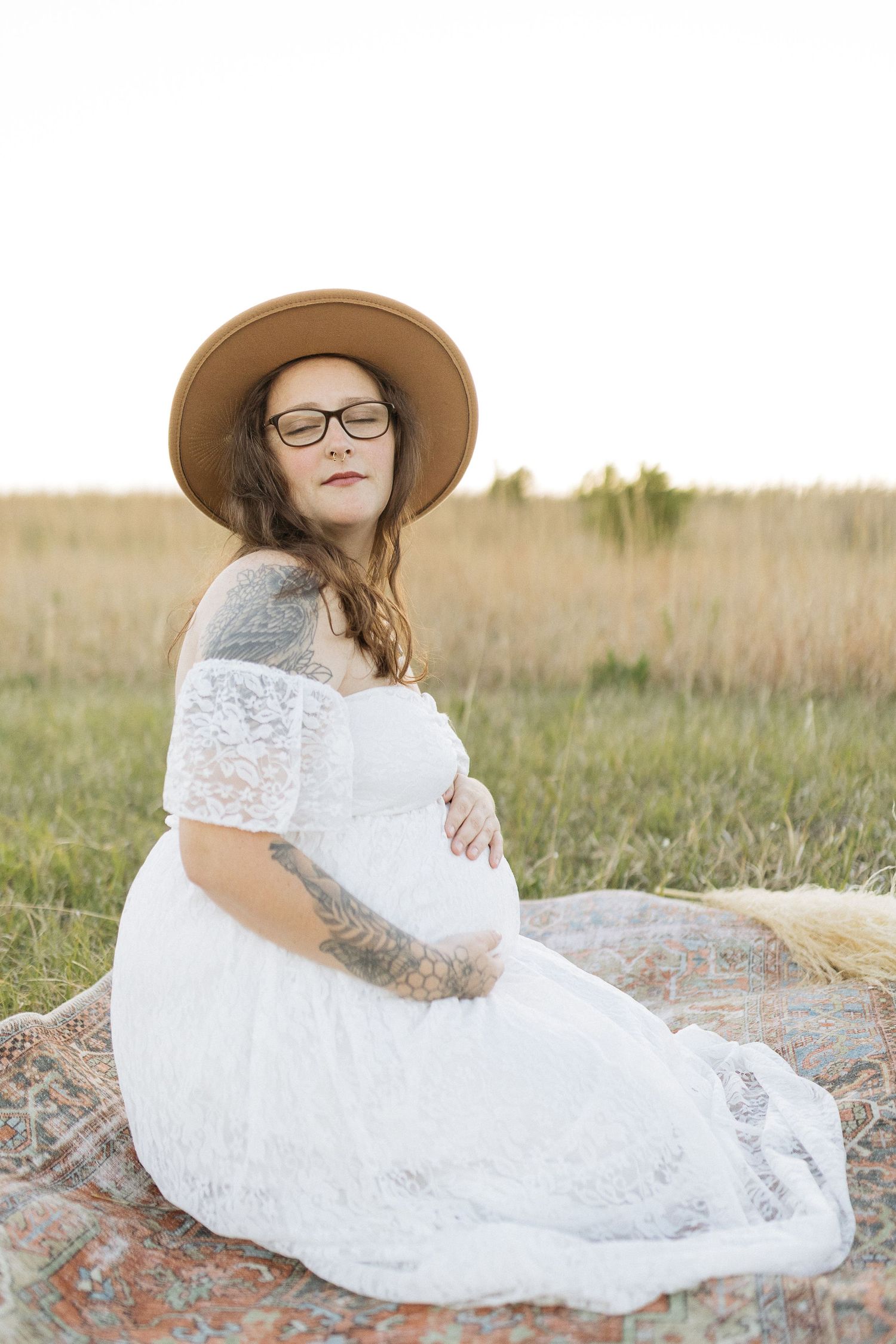 A maternity portrait in a field featuring a white lace dress and brown wide-brim hat.