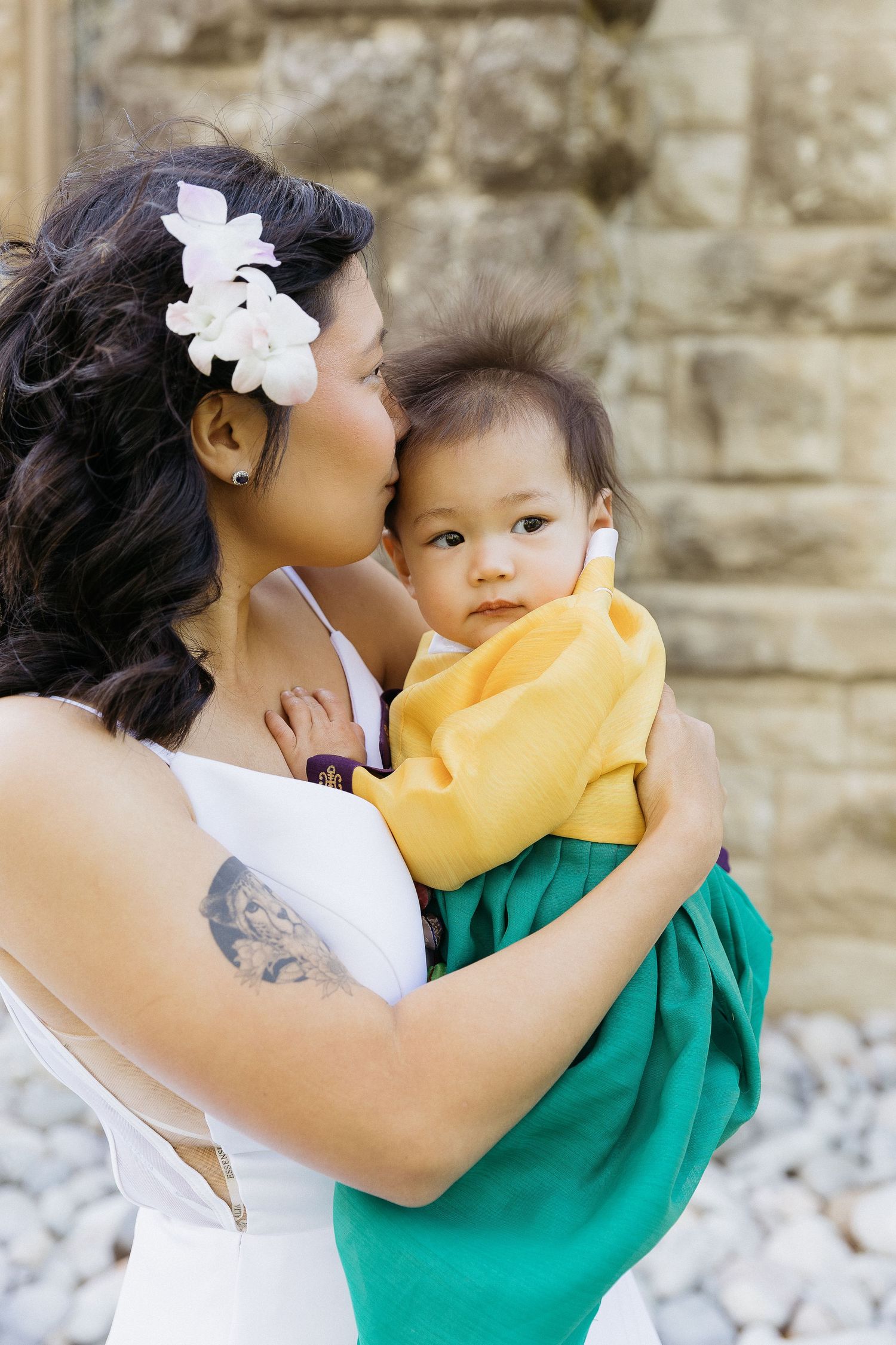 A tender family moment as a parent in a white dress holds a baby wearing yellow and green against a stone wall background.