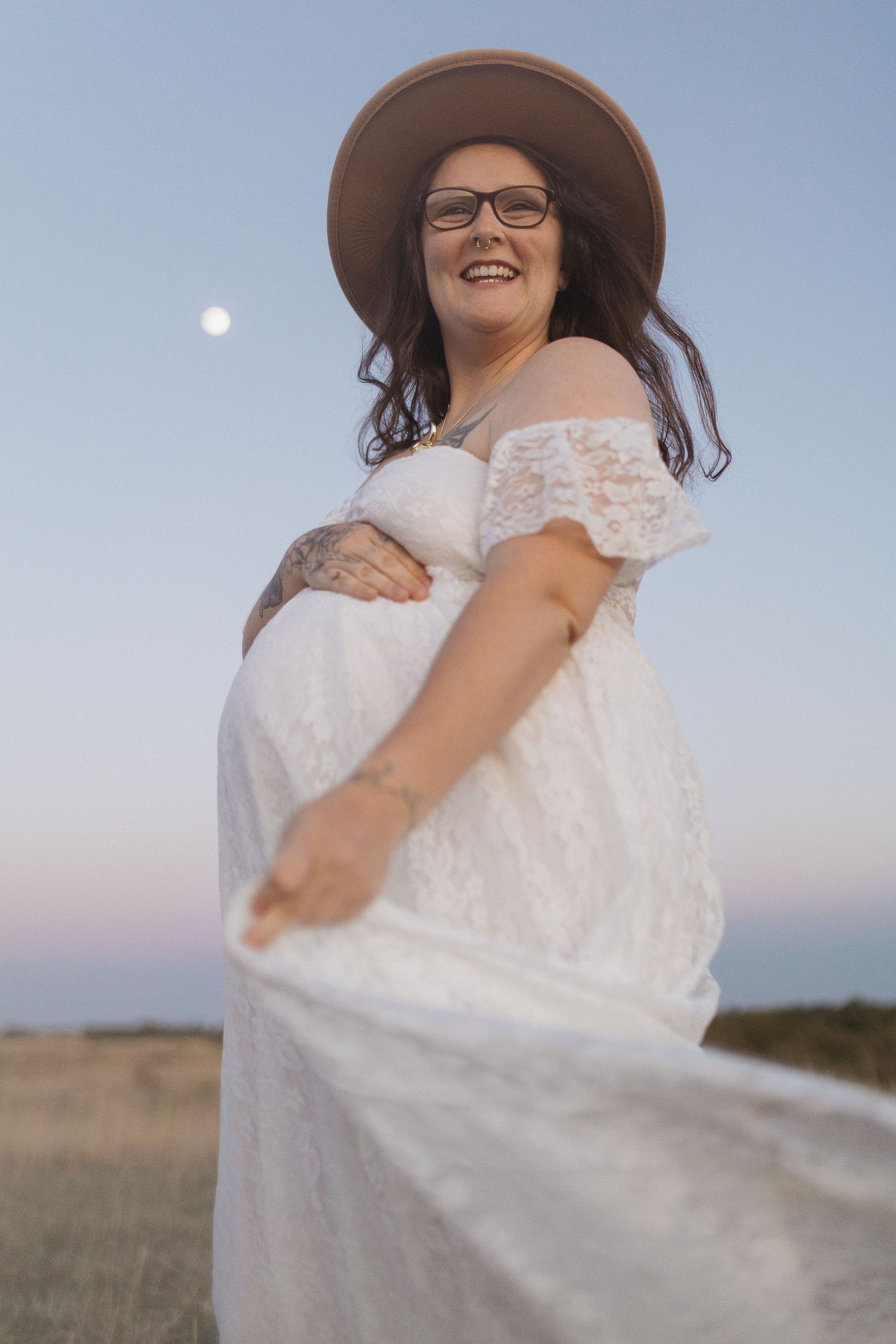 A joyful maternity portrait featuring a white lace dress and brown hat against the evening sky.