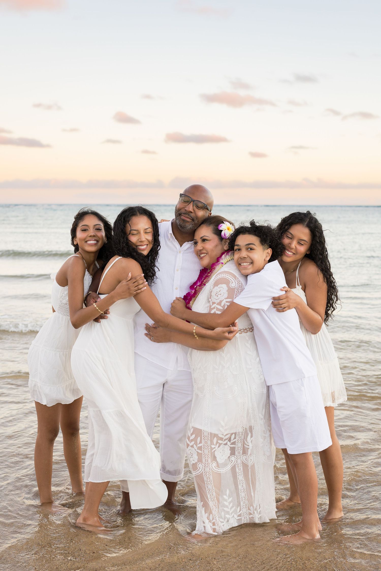 A joyful family group embraces on a beach at sunset wearing coordinated white outfits.