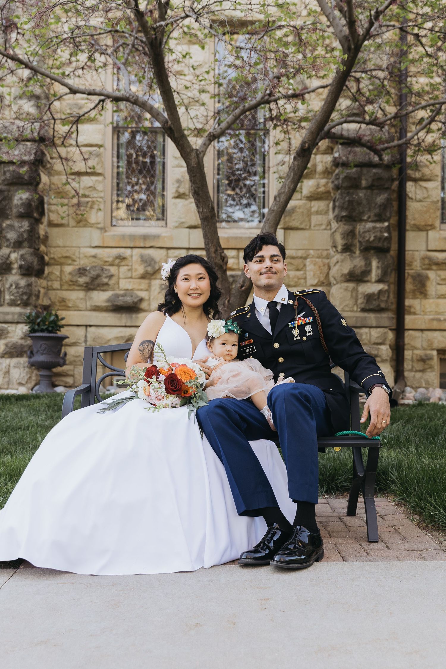 A military wedding couple sits on a bench with their baby in front of a stone church.