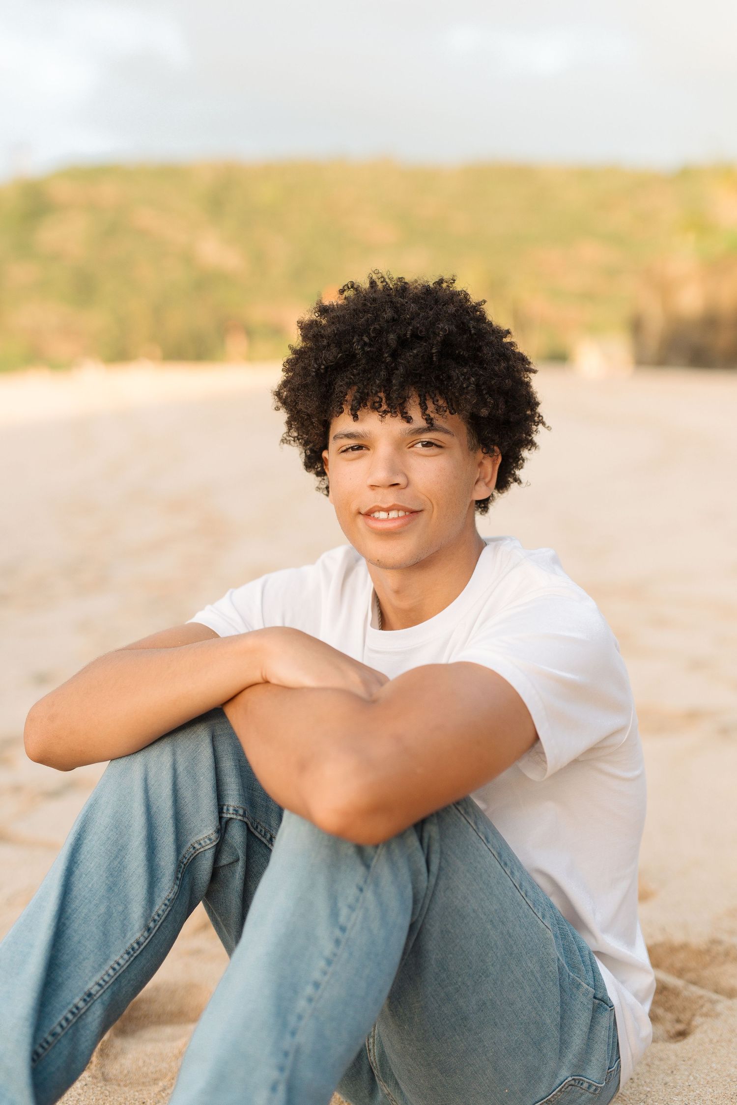 Warm sunset portrait of someone sitting on beach sand wearing white t-shirt and jeans.waimea bay senior session north shore oahu hawaii