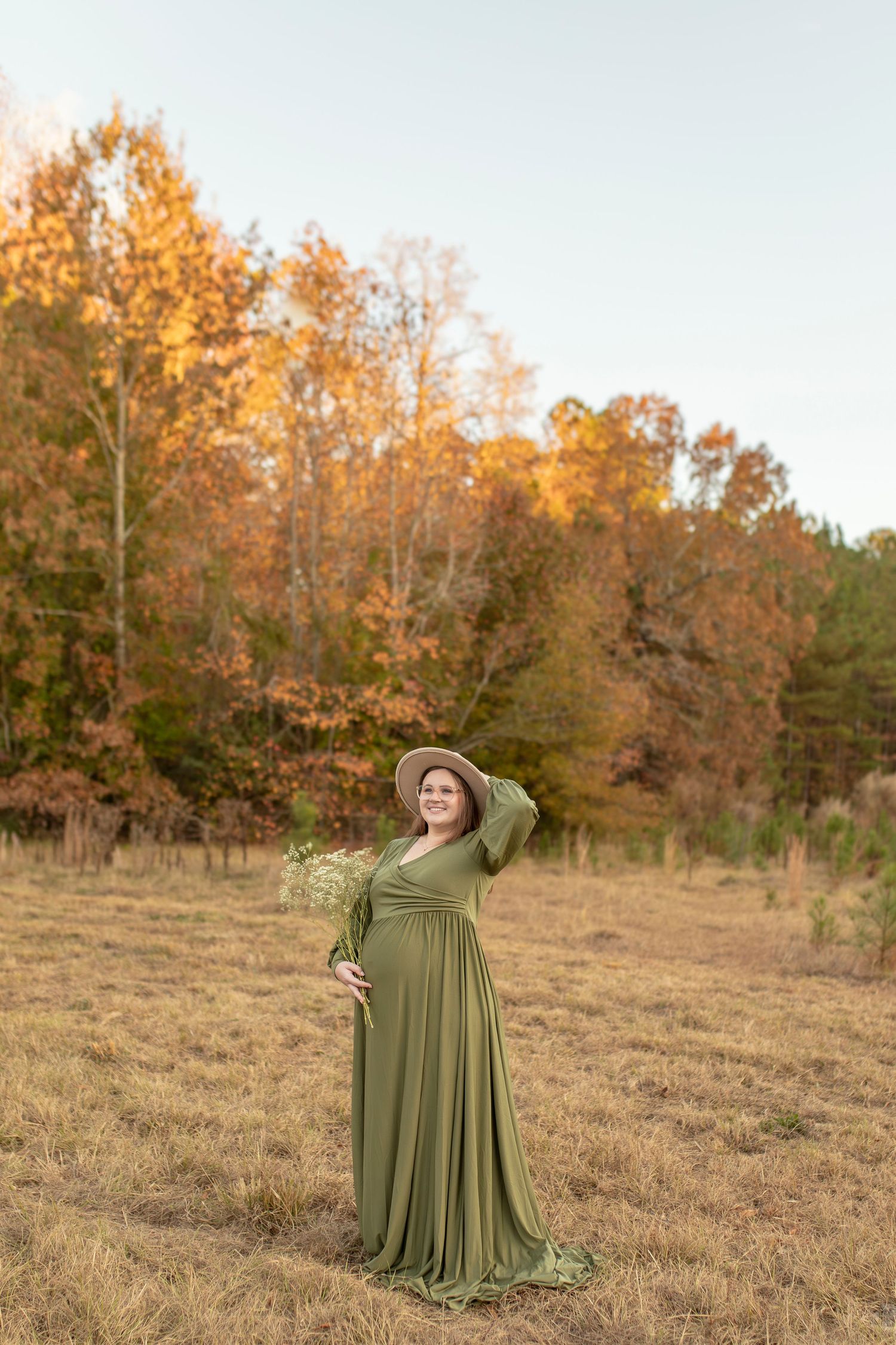Woman in flowing olive green dress poses gracefully in autumn field at sunset.