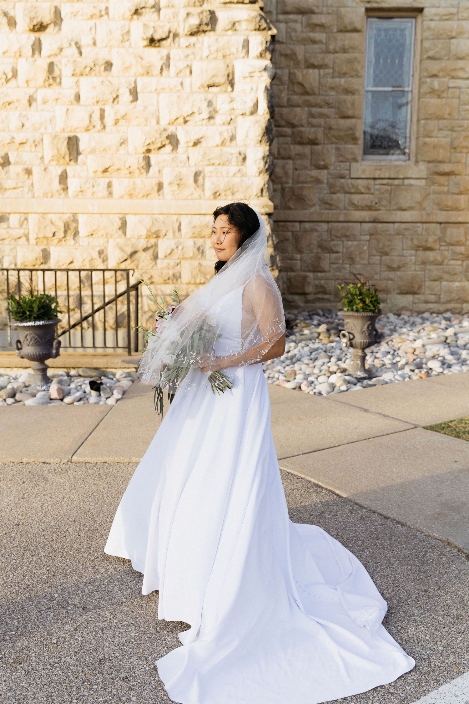 A bride in a flowing white dress and veil poses against a stone church wall.