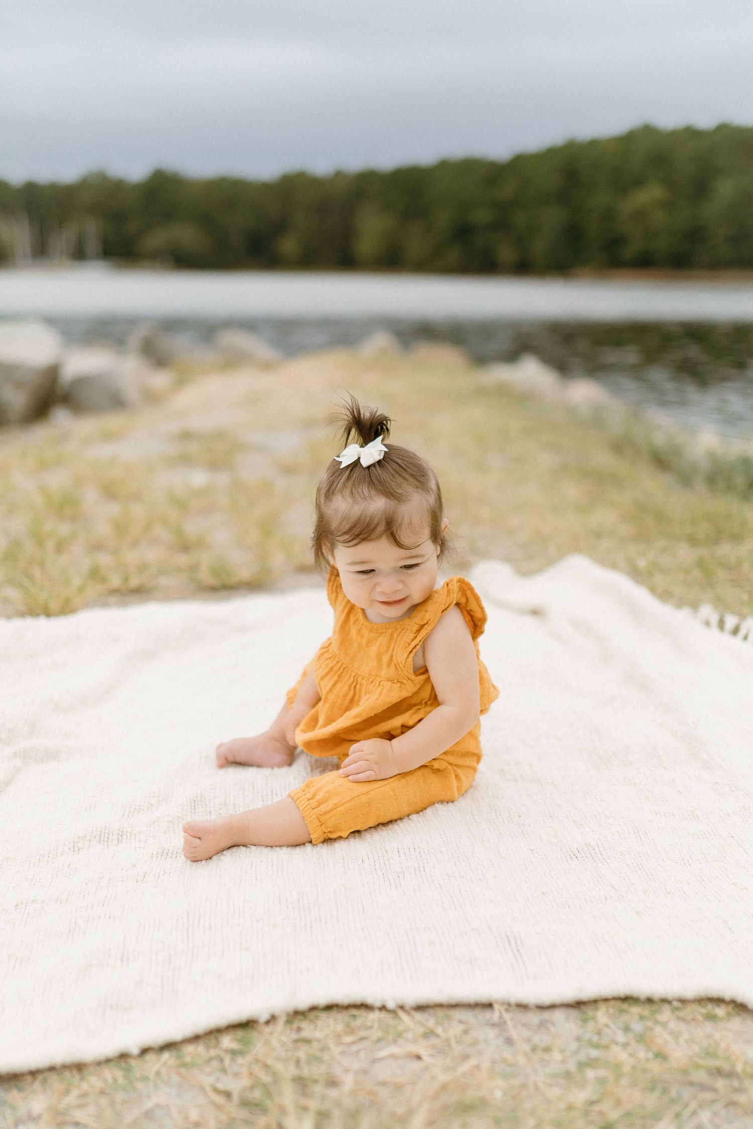 Small child in yellow romper sits on blanket by lakeside on sunny day.