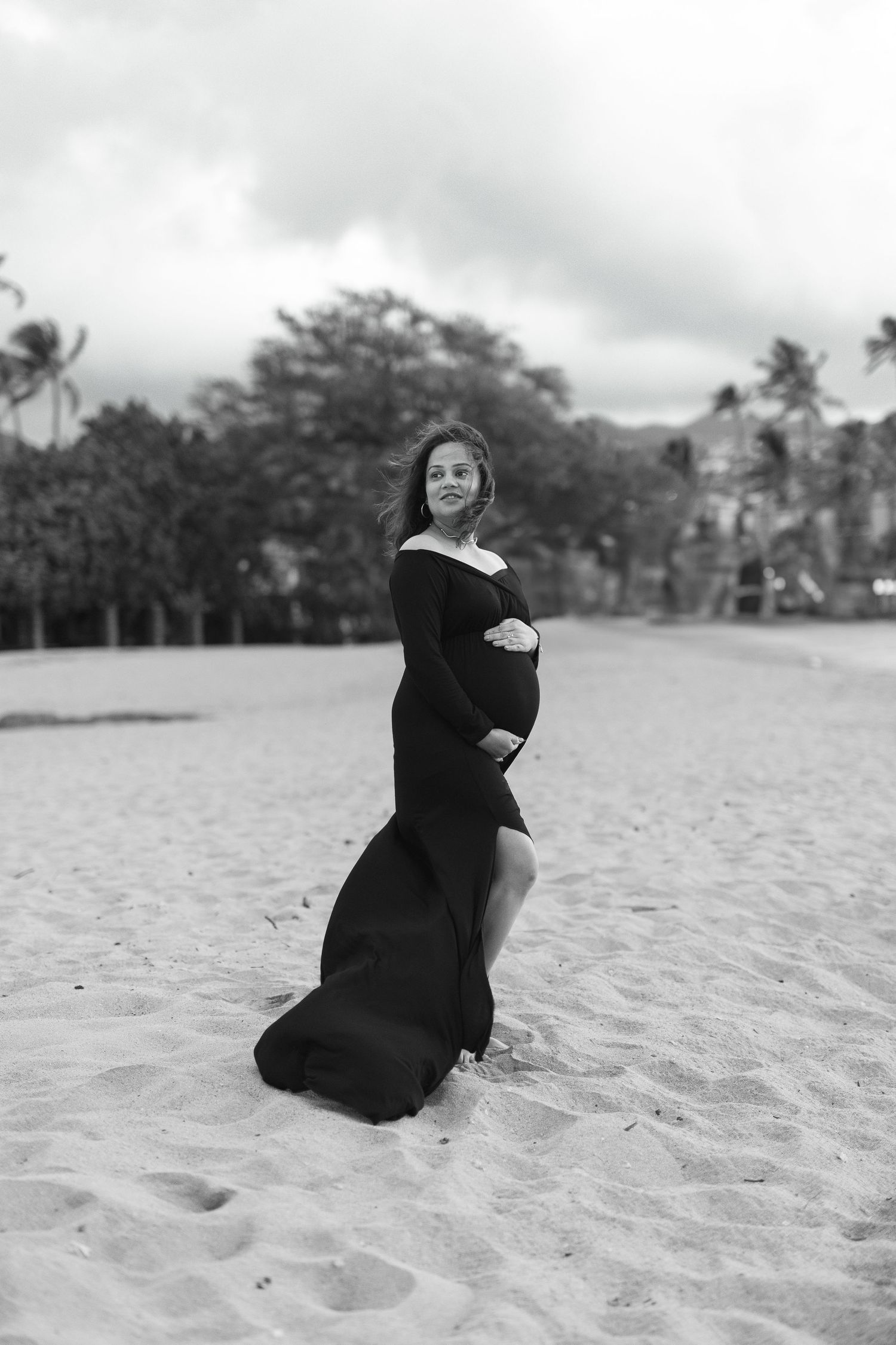 A black and white photo of someone in an elegant off-shoulder black dress posing on a sandy beach with palm trees.