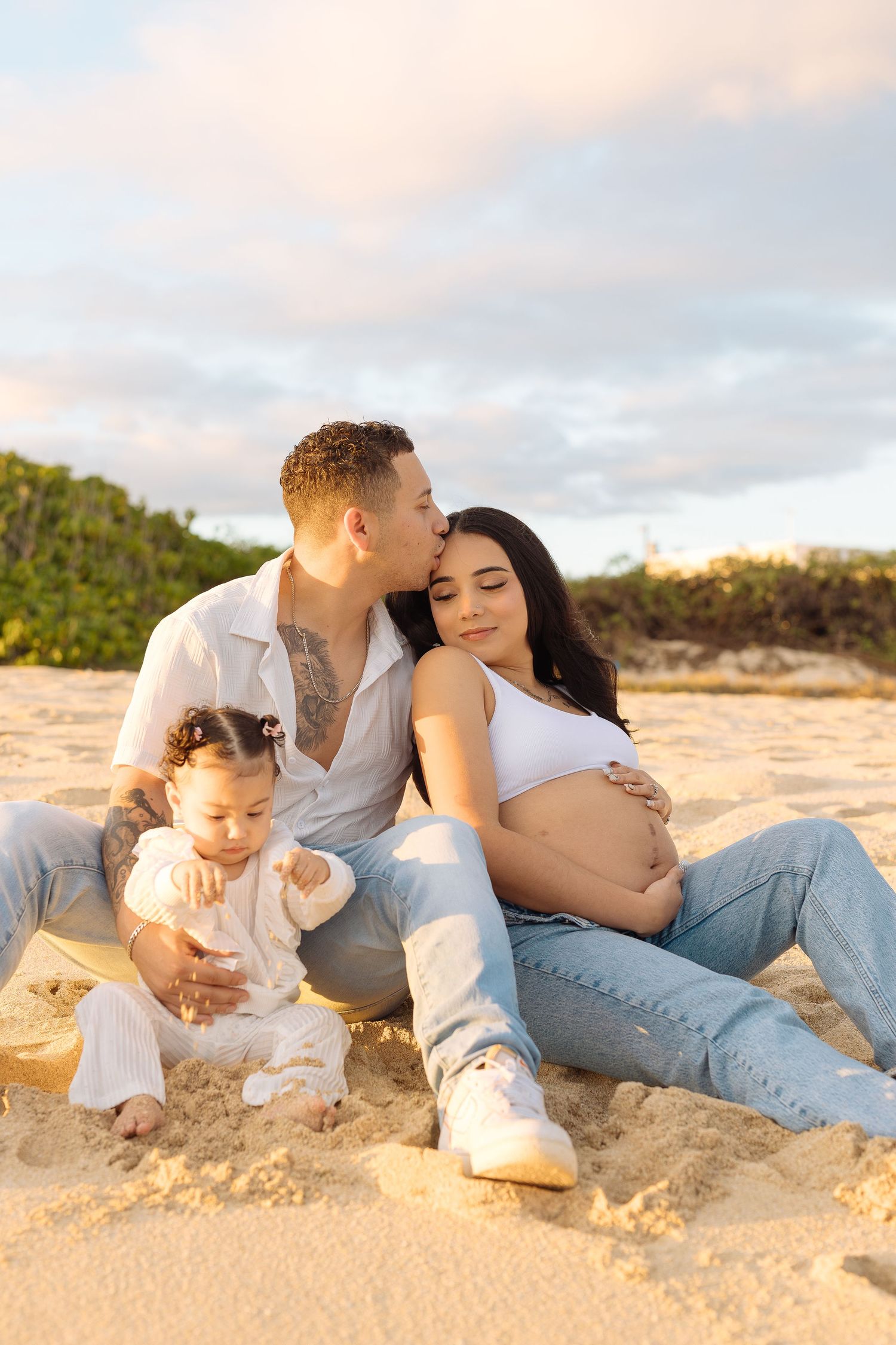A family sits together on a sandy beach during sunset for a maternity photo session.