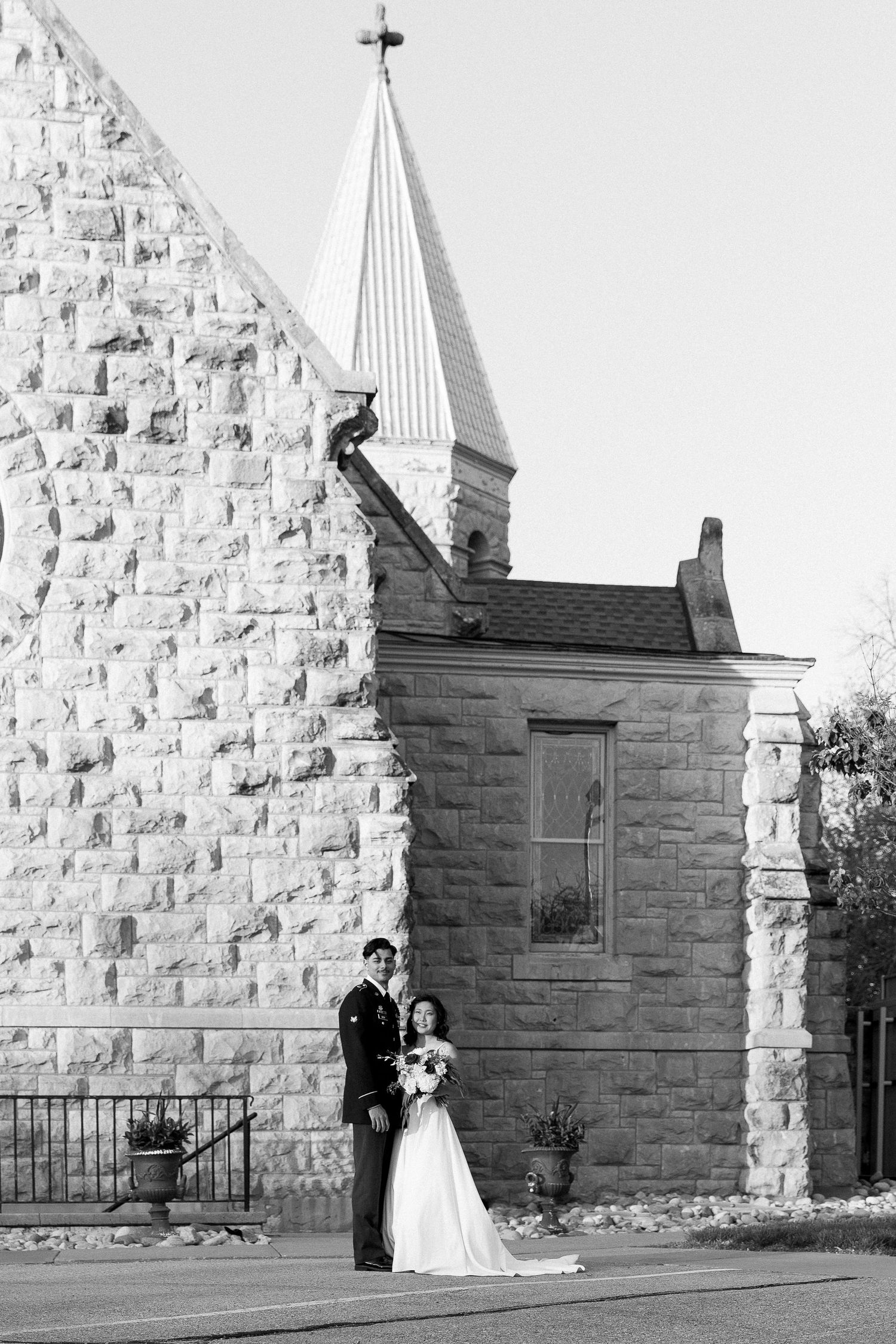A black and white wedding photo featuring a couple beside a stone church with a tall spire.