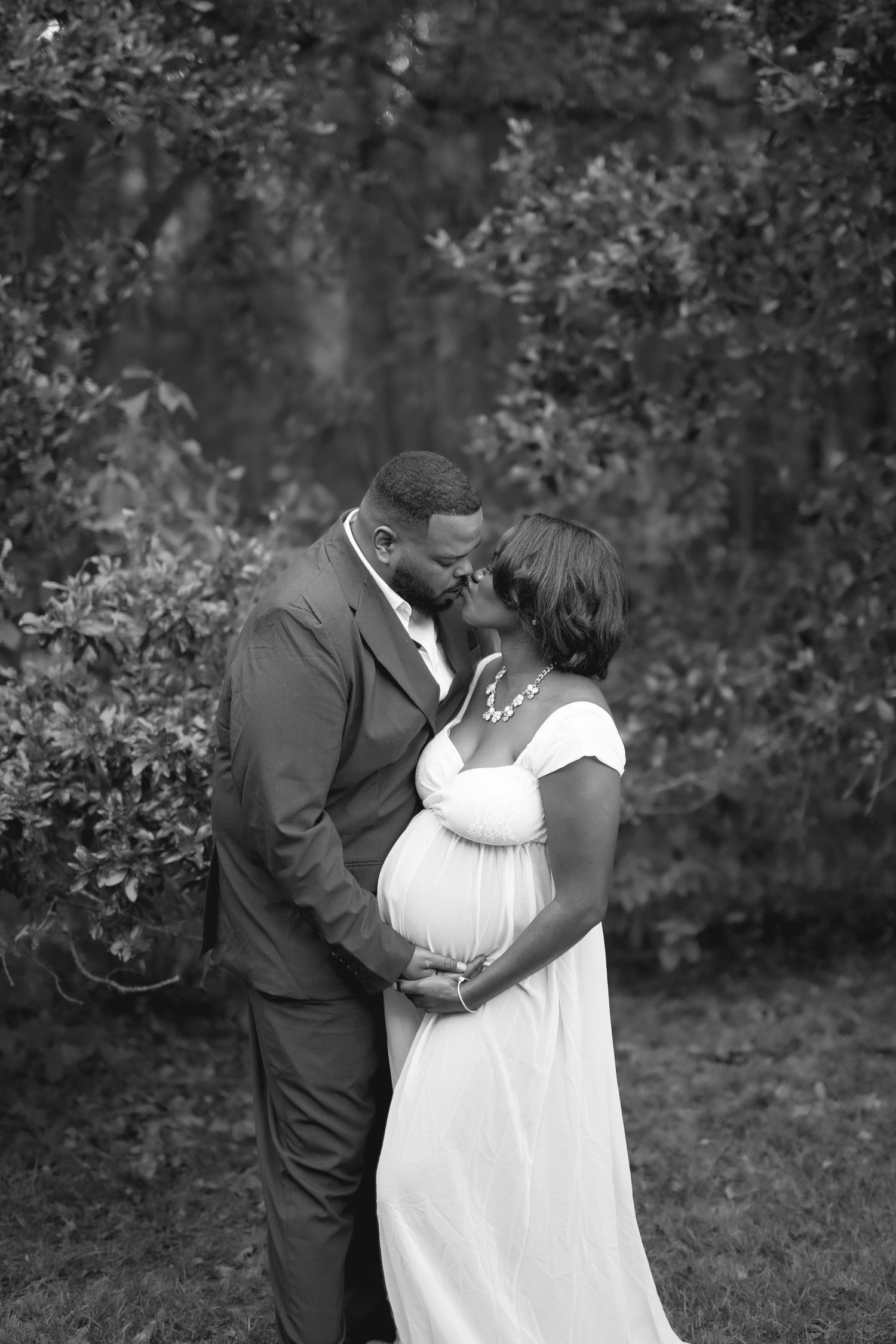 Intimate black and white maternity portrait of a couple sharing a tender moment in a garden setting.