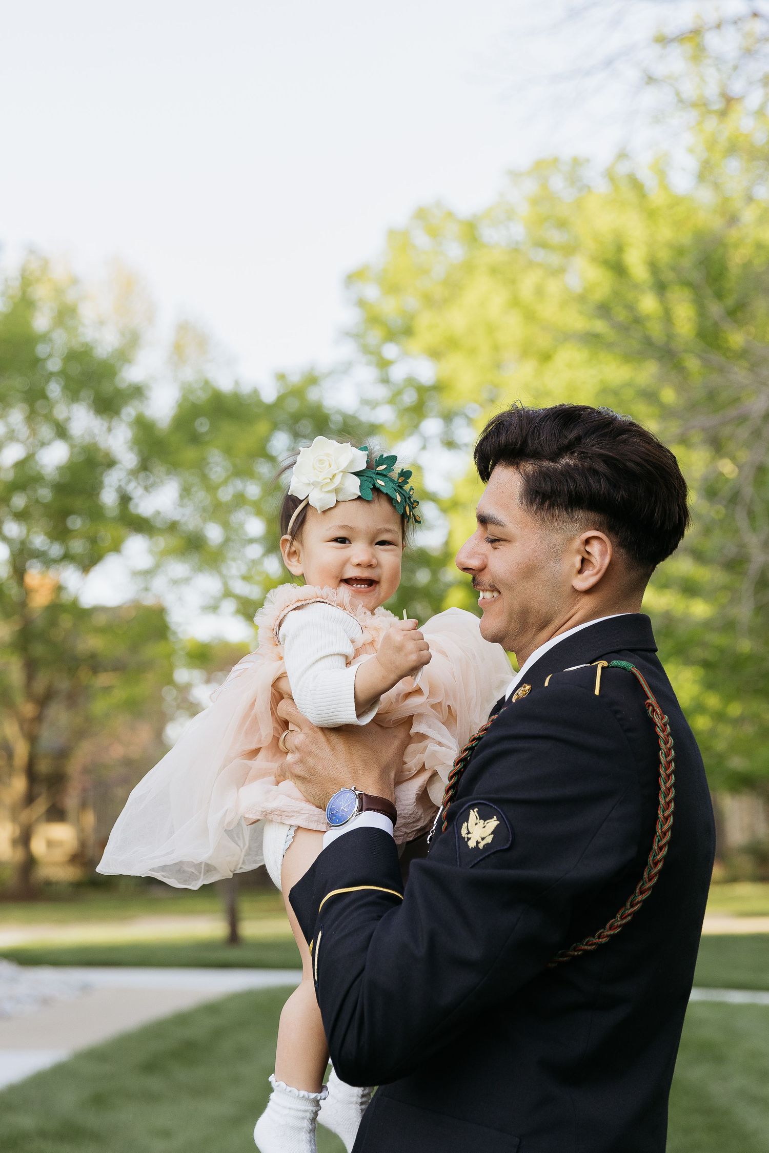 Military servicemember in dress uniform holds smiling baby in pink dress outdoors.