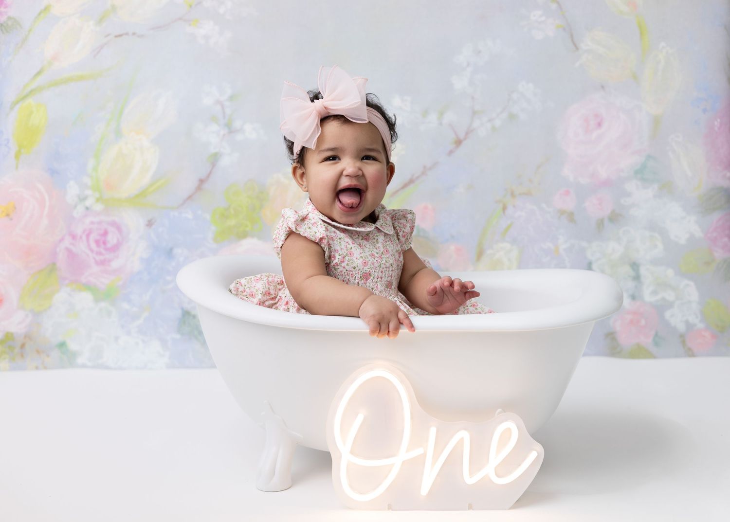 A one-year-old girl smiles happily while sitting in a bathtub, celebrating her first birthday with bubbles and fun decorations.