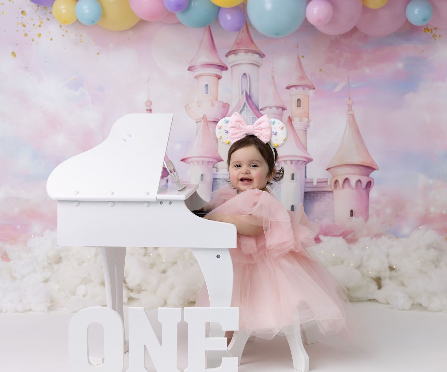 A one-year-old girl in a Disney-themed outfit plays on a toy piano, surrounded by colorful decorations.
