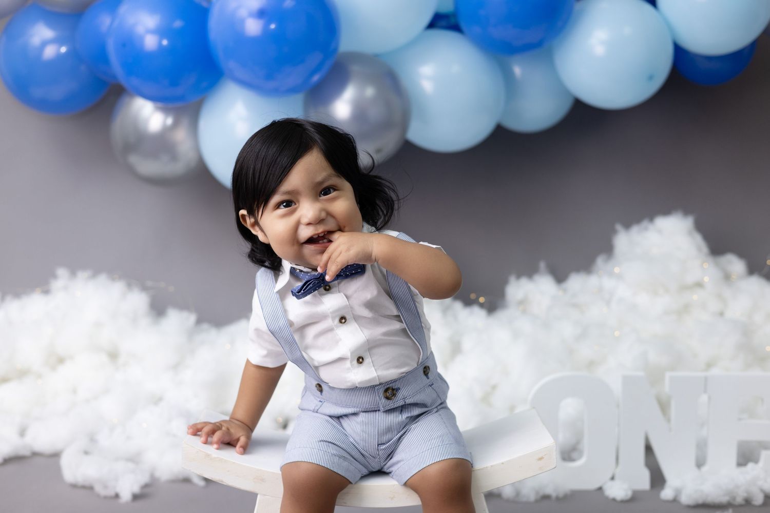 A one-year-old birthday boy sits on a chair, smiling happily during his first birthday photo session.