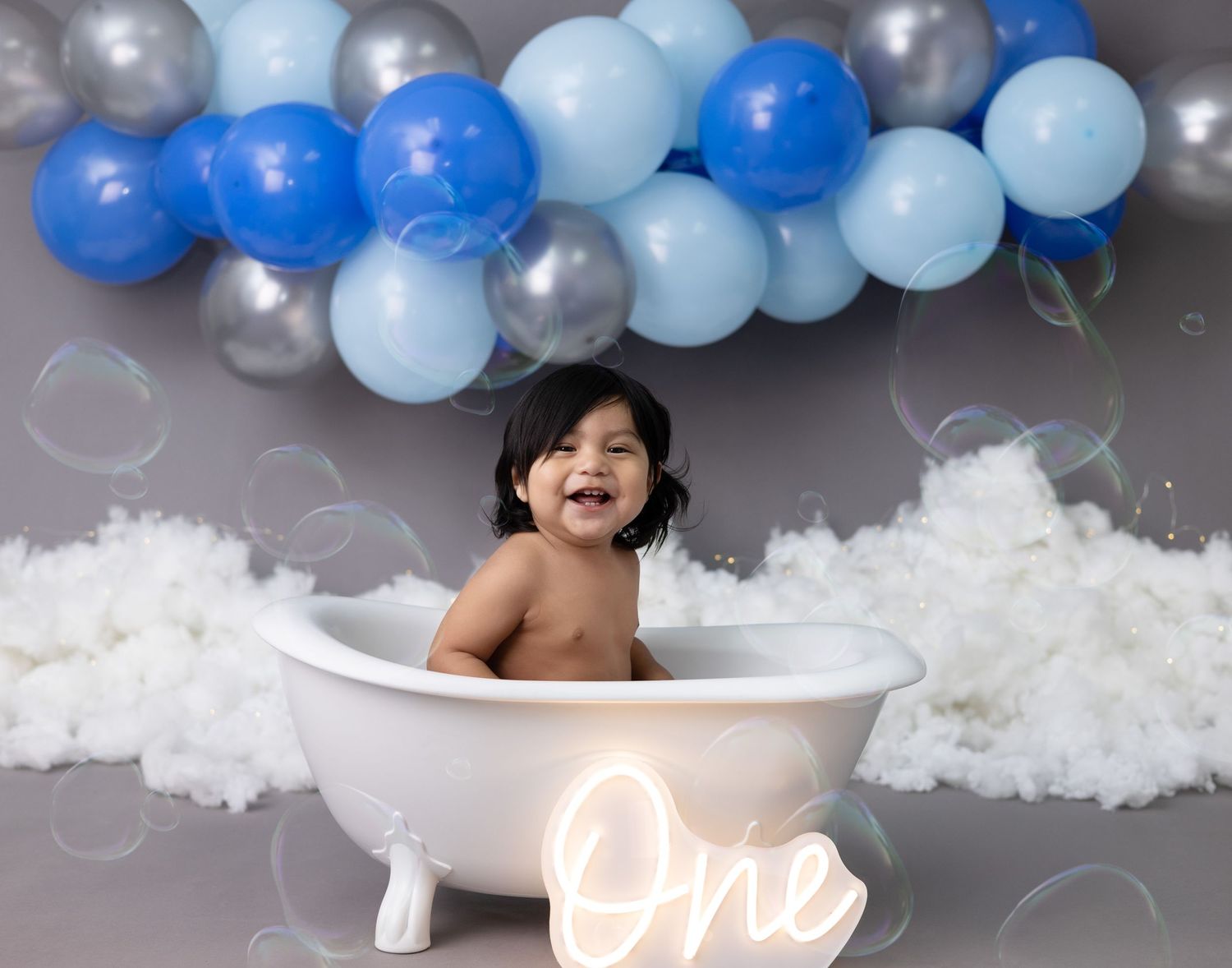 A one-year-old boy sits in a bathtub, smiling and splashing, celebrating his first birthday.