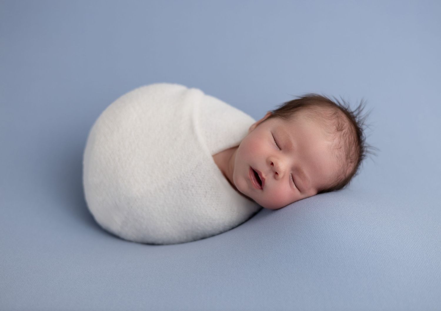 A newborn baby boy, swaddled in a soft, neutral-toned blanket, sleeps peacefully with his tiny hands tucked in.
